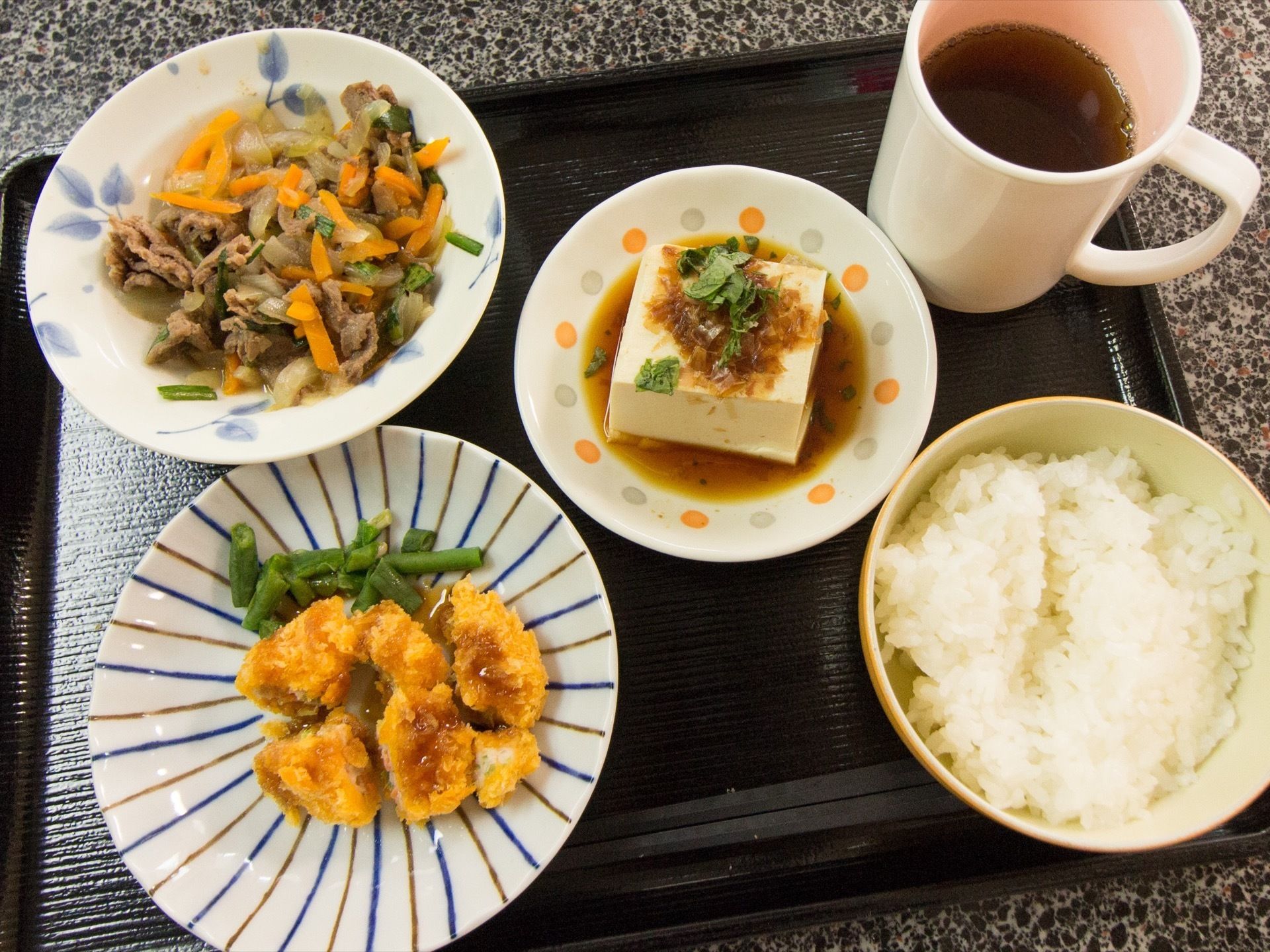 黒いお盆の上に、お茶碗に盛られた白いご飯と揚げ物、煮物のボウルと冷奴が乗っている。マグカップにはお茶がそそがれている。