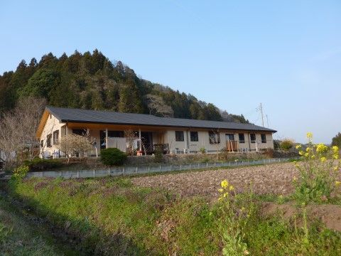山の前に建つ三角屋根の平屋で、窓も多く設けられている。外壁は白、屋根は黒い横長の家屋。家の前には畑があり、四方は自然に囲まれている。