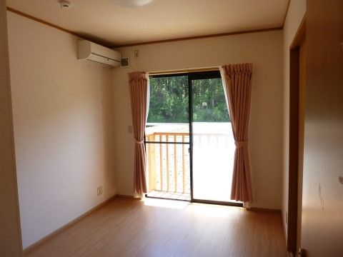 エアコンが付いた、白い壁の居室である。大きな窓には白いレースとベージュ系のカーテンがあり、窓の外には緑の自然が広がっている。