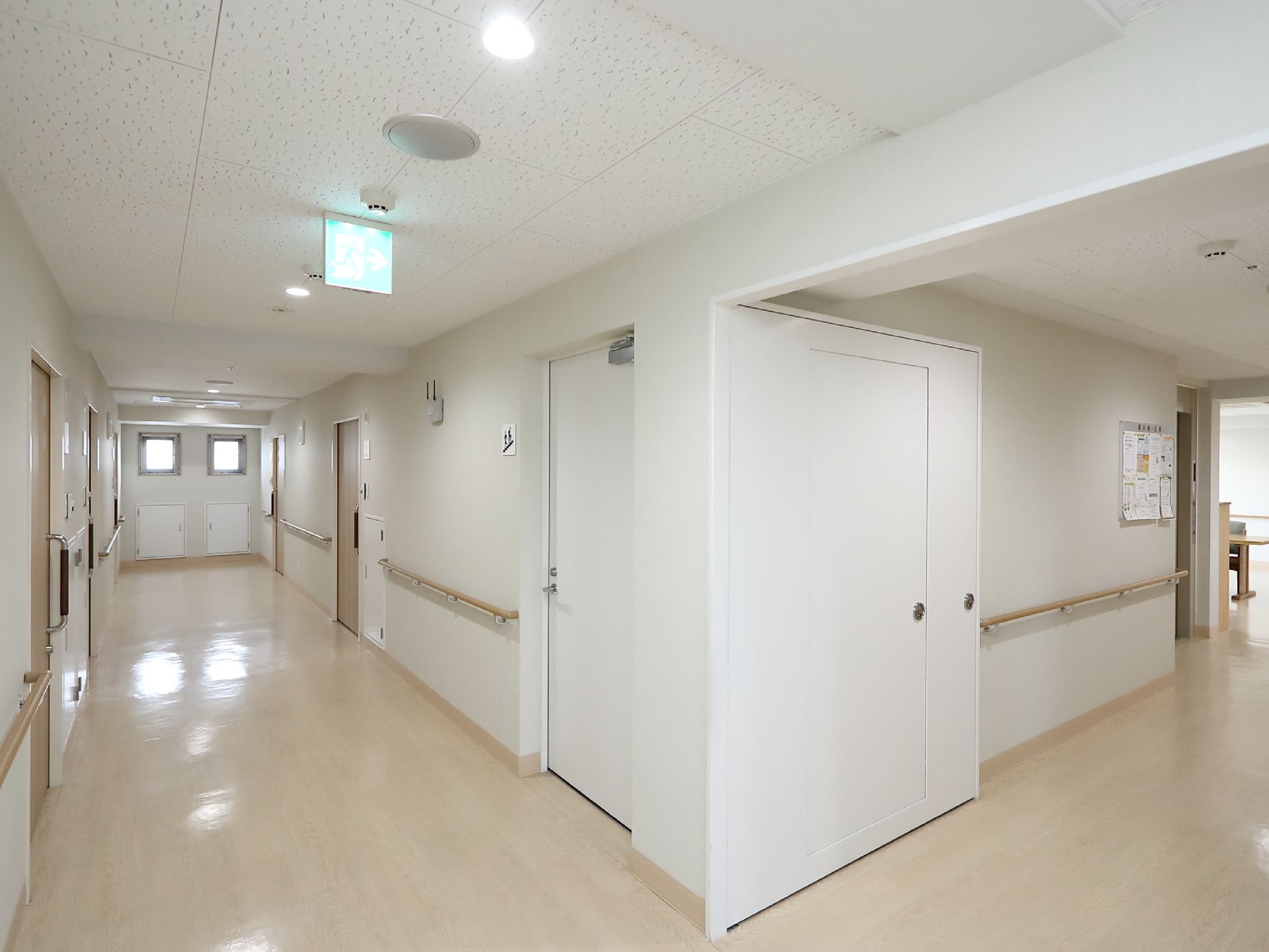 白い壁の通路である。床は淡いベージュで突き当たりに四角い小窓が２つある。壁には手すりが取りつけられている。