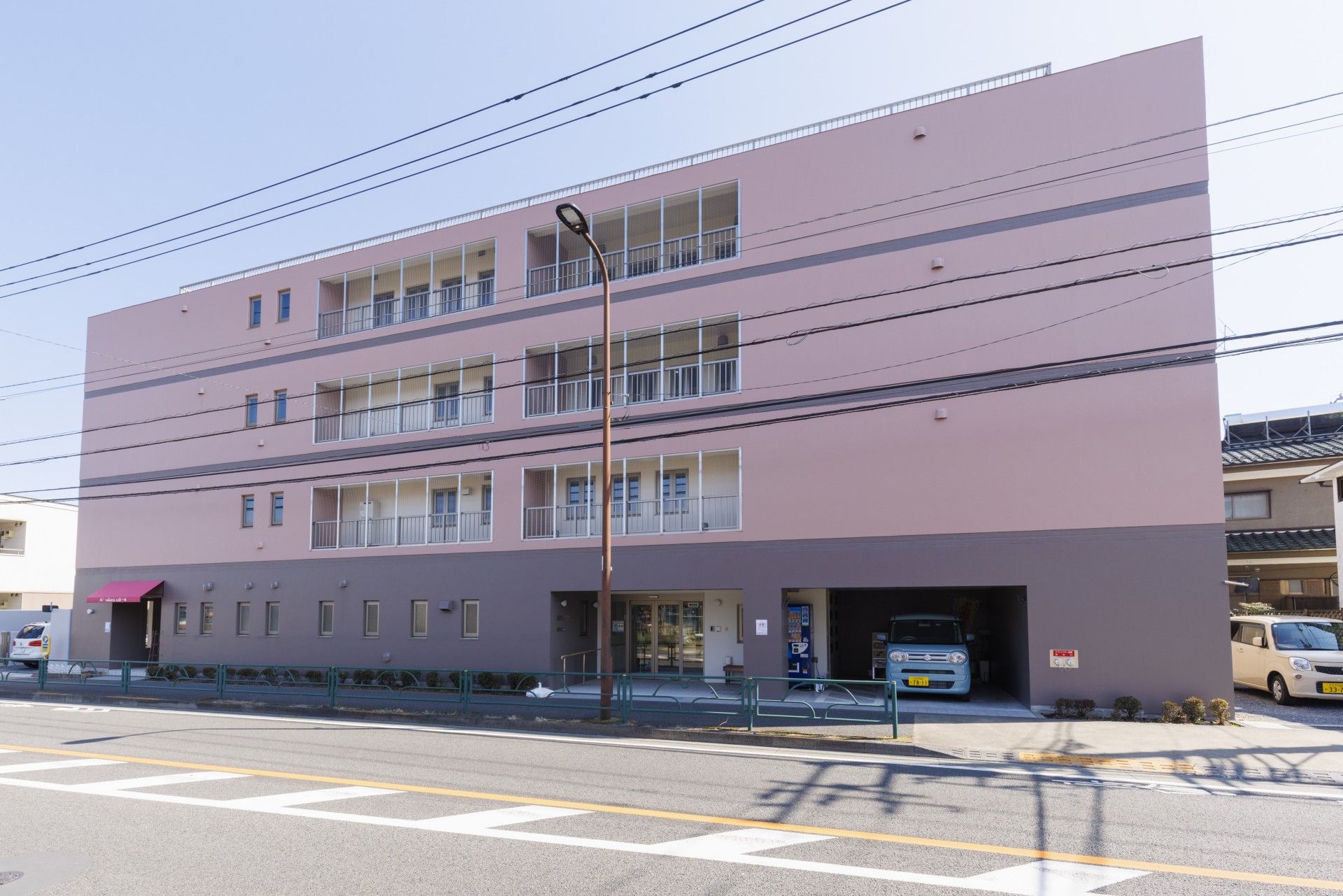 駐車場に車が１台止まっていて、その横に青い自動販売機が設置されている。建物の周辺には住宅があるのが見える。