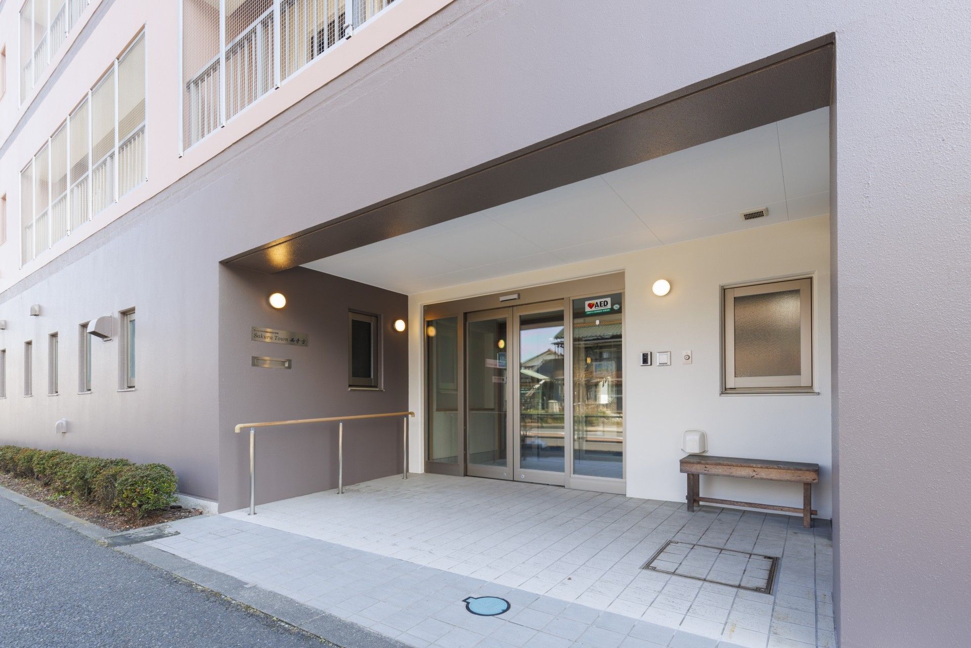 ガラスの自動ドアが設置されている。壁には手すりが取りつけられていて、ドアの横に木製のベンチが置かれている。