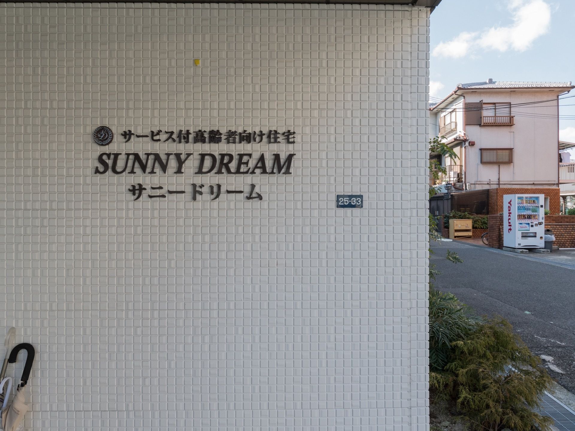 屋根がある玄関前のスペースになっており、雨の日も濡れずに施設内に入ることができる。施設名が表示されており、文字はダークブラウンである。