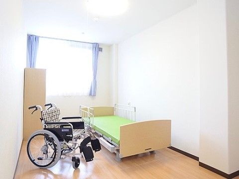 居室には大きな窓があり、フローリングの床には介護用ベッドと収納家具が置かれている。壁と天井の色は白色系で、照明設備が完備されている。