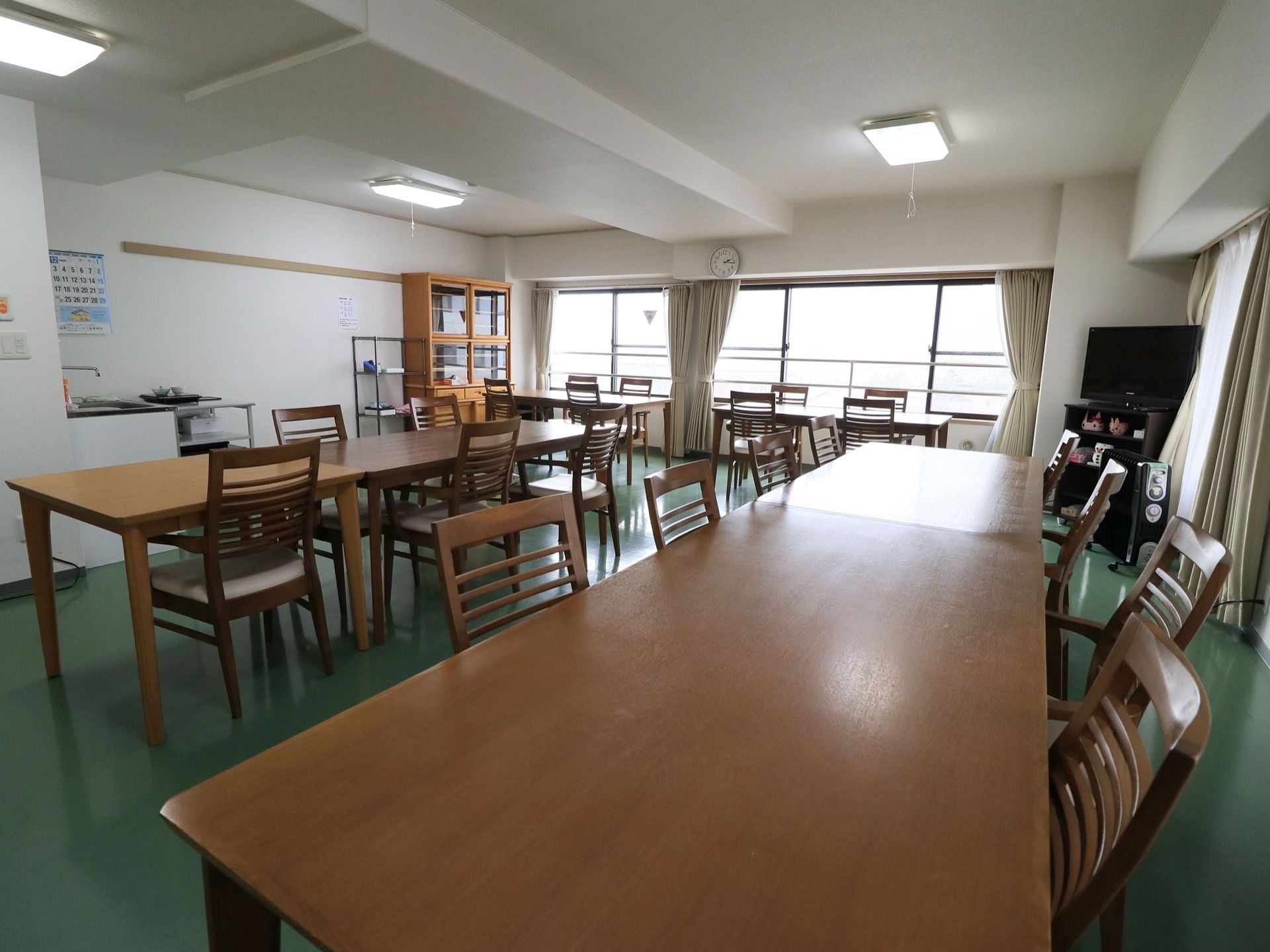 食堂には茶色いテーブルと椅子が複数並べられている。食器棚もあり、隣にはメタルラックが置かれている。テレビも1台ある。