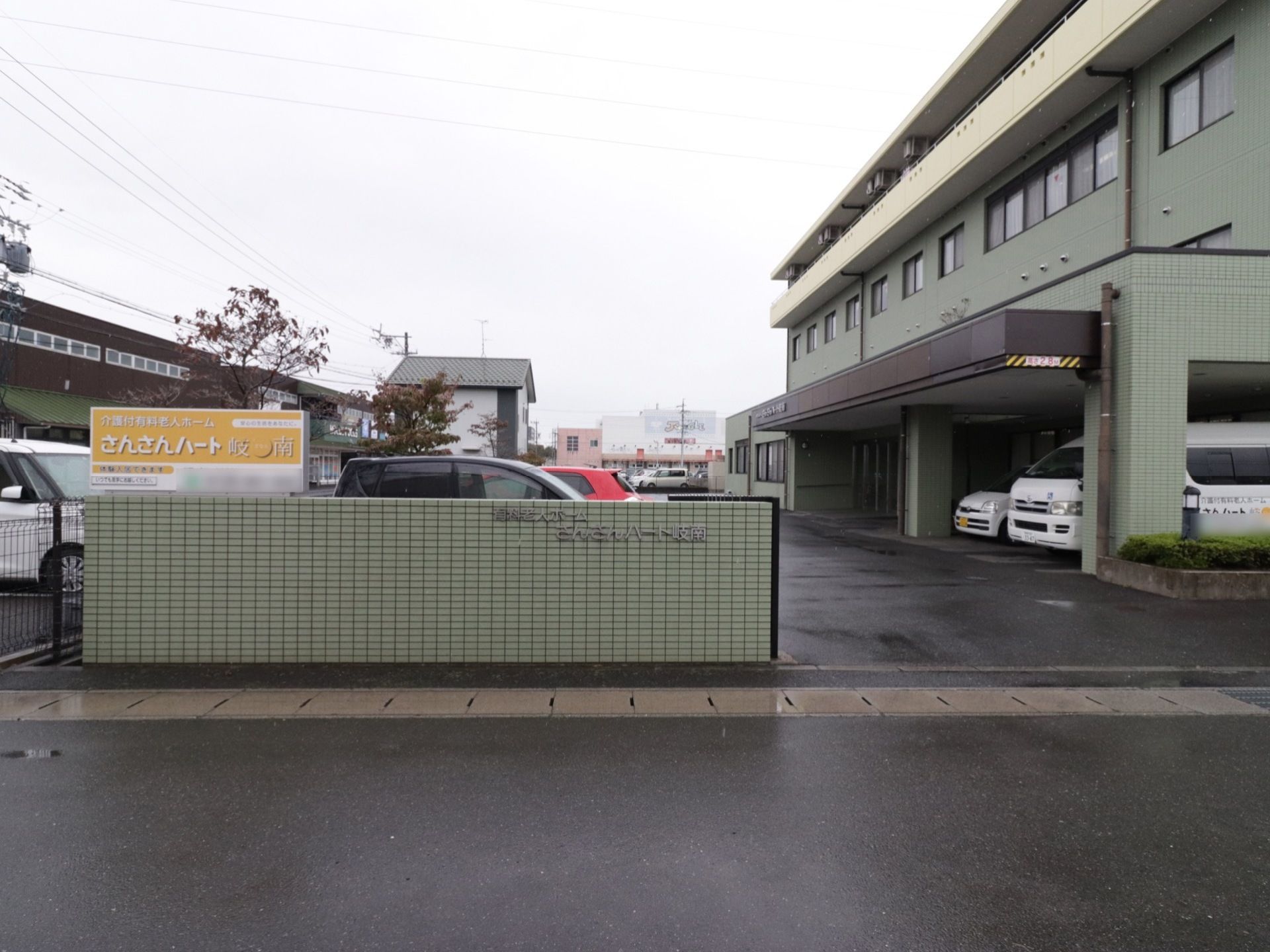モスグリーンの壁の左端に、黄色い看板が取り付けられている。その向こうには駐車場があり、数台の車が停まっている。