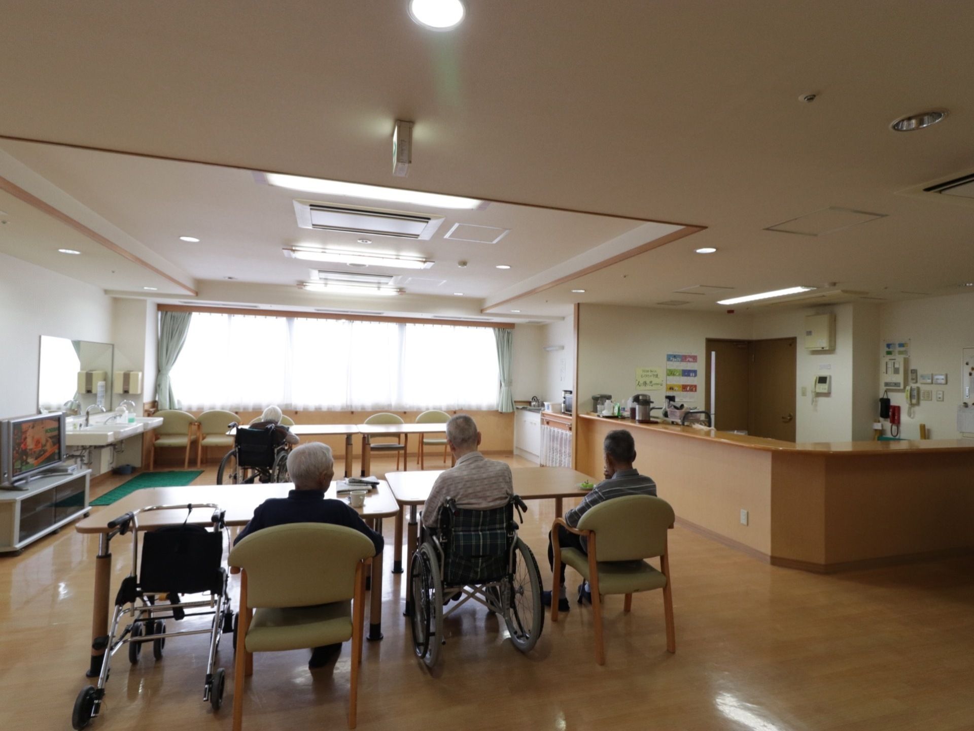 手前のテーブルには三名、奥のテーブルには一名の入居者様が座っている。窓の右横には調理台があり、ヤカン等が置いてある。