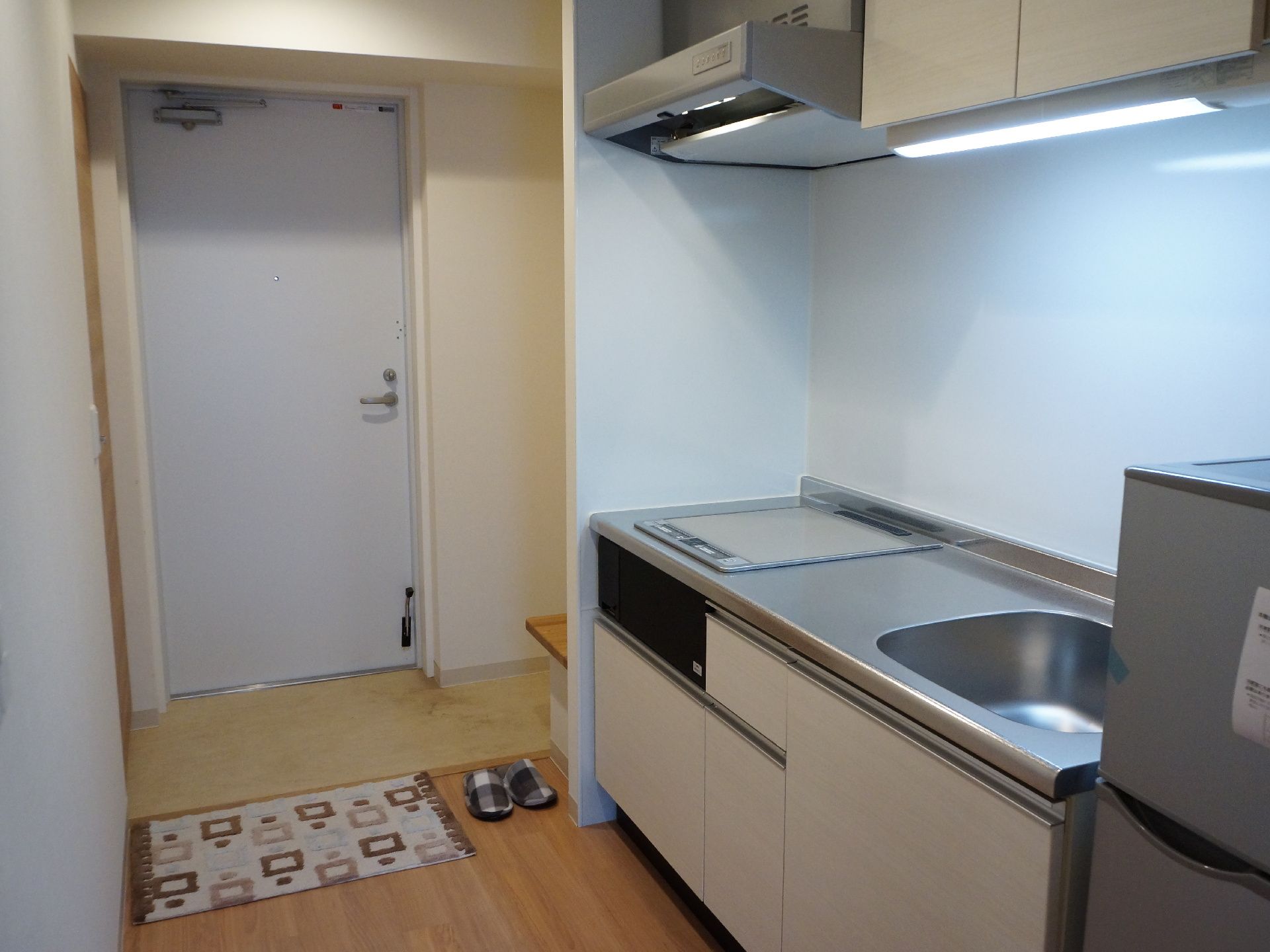 居室の入り口とキッチン。玄関には腰掛と手摺りを設置。また大き目の収納も用意されている。キッチンはIHクッキングヒーターで安全にご利用できる。