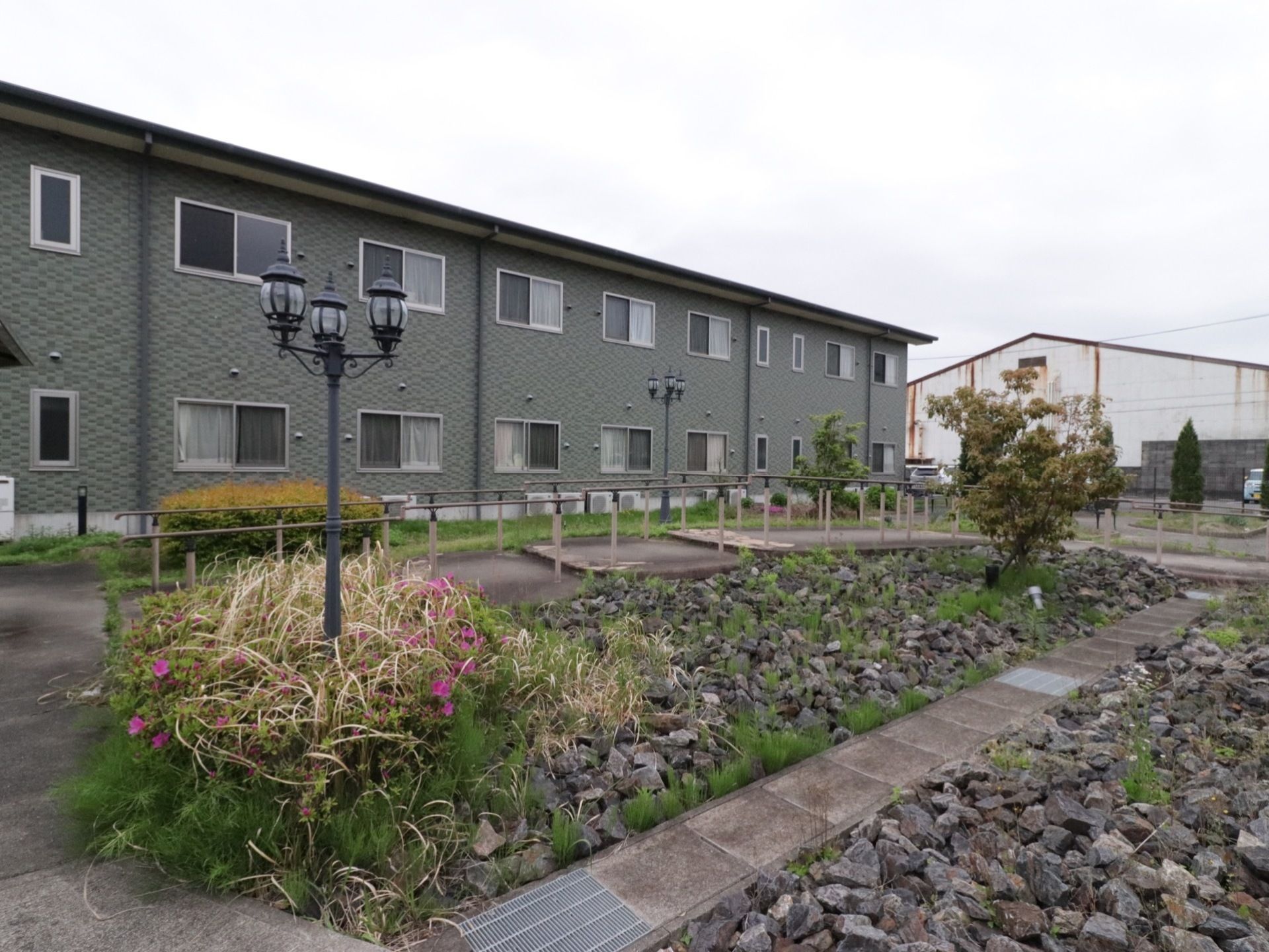 向こう側に2階建ての建物があり、その前に庭が広がっている。石が敷き詰められたスペースに木々が植えられ、街頭も立っている。