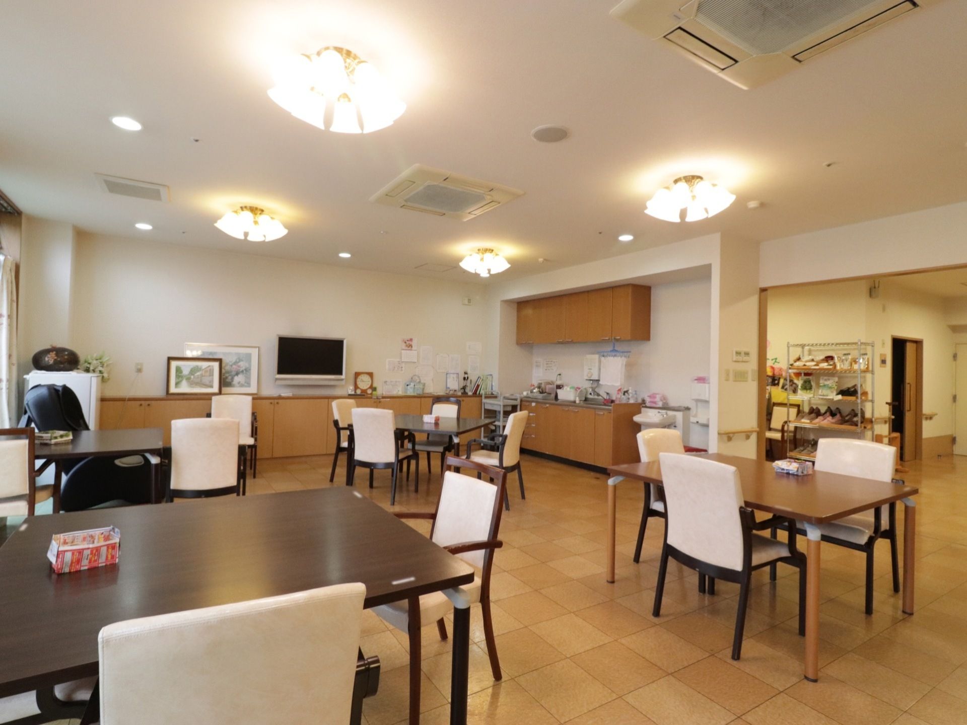 室内に、テーブルセットが4つある。天井には、おしゃれな照明が付いている。部屋の奥にはテレビが設置されており、右側にはキッチンがある。