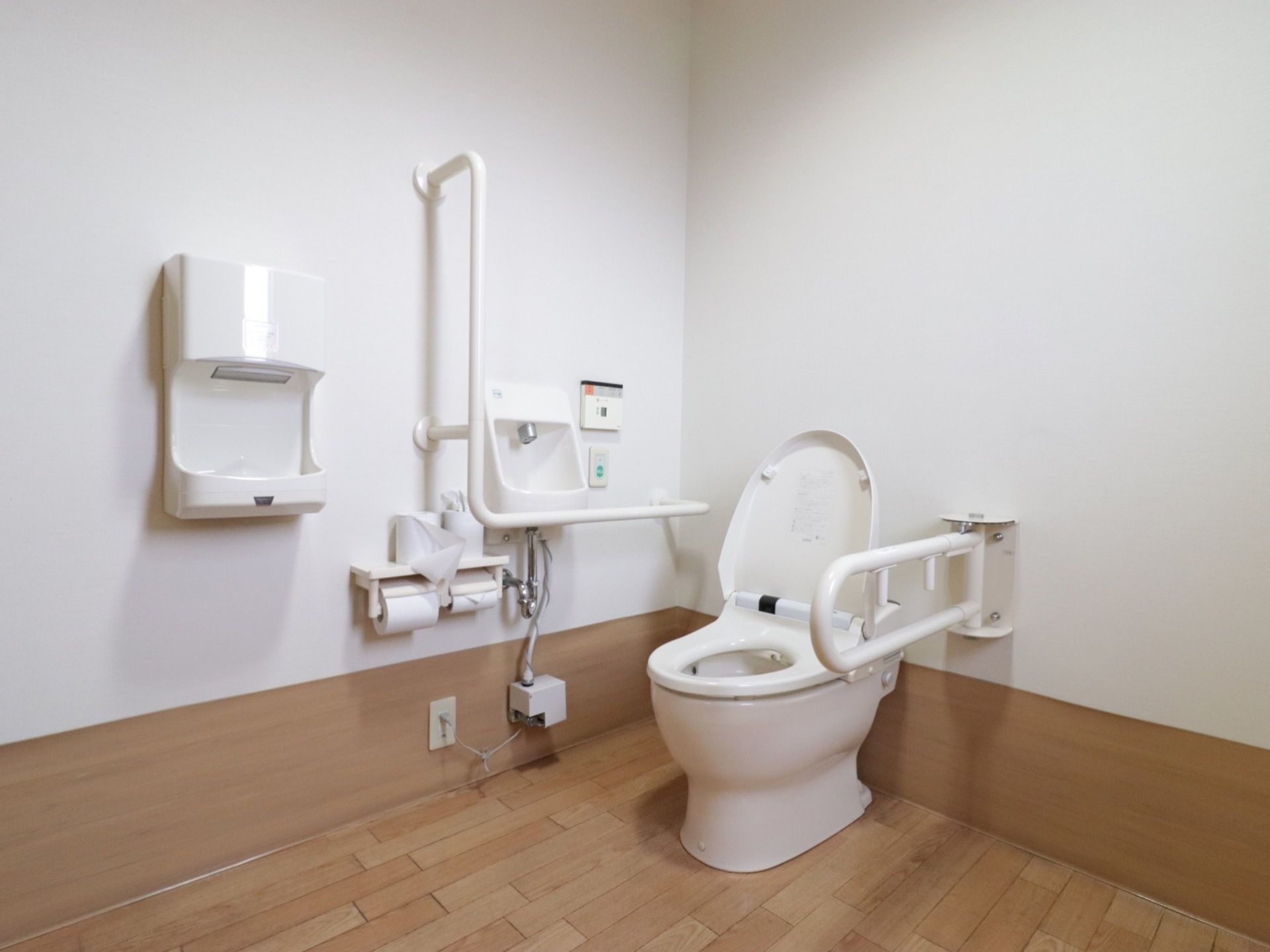 トイレ個室内の様子。温水洗浄便座付きトイレで、壁に操作盤がある。壁に手すりや手洗い場、ナースコールが設置されている。