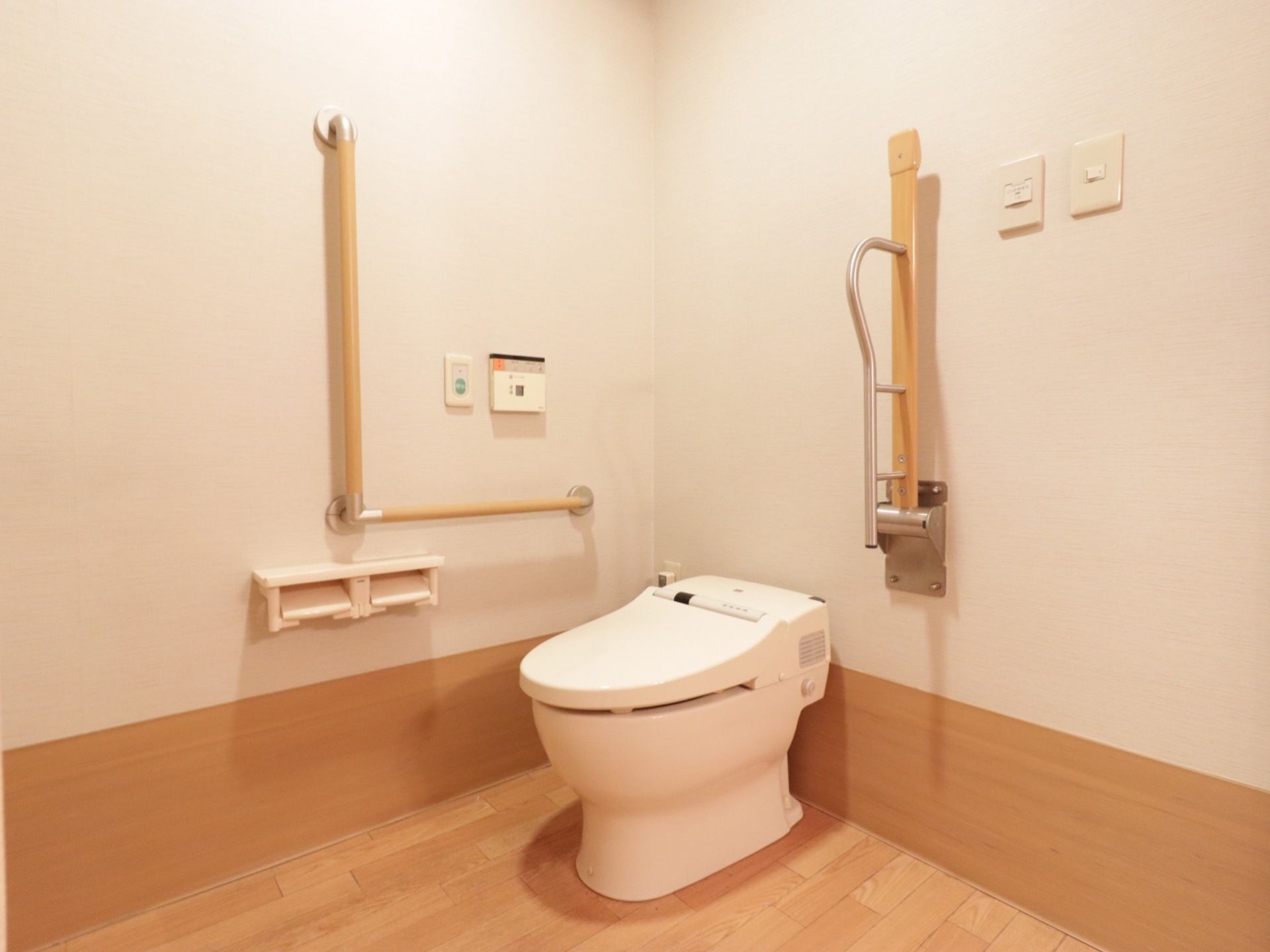 トイレ個室内の様子。角に洋式便器がある。壁にL字型の手すりがあり、操作盤とナースコールが設置されている。