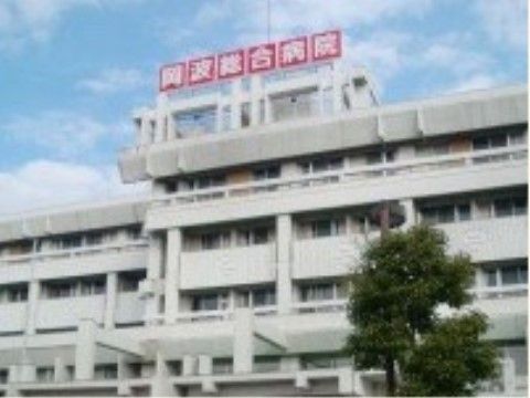 白い外壁の病院である。屋上には、施設名が書かれた赤い看板が取り付けられている。手前に大きな木が植えられている。