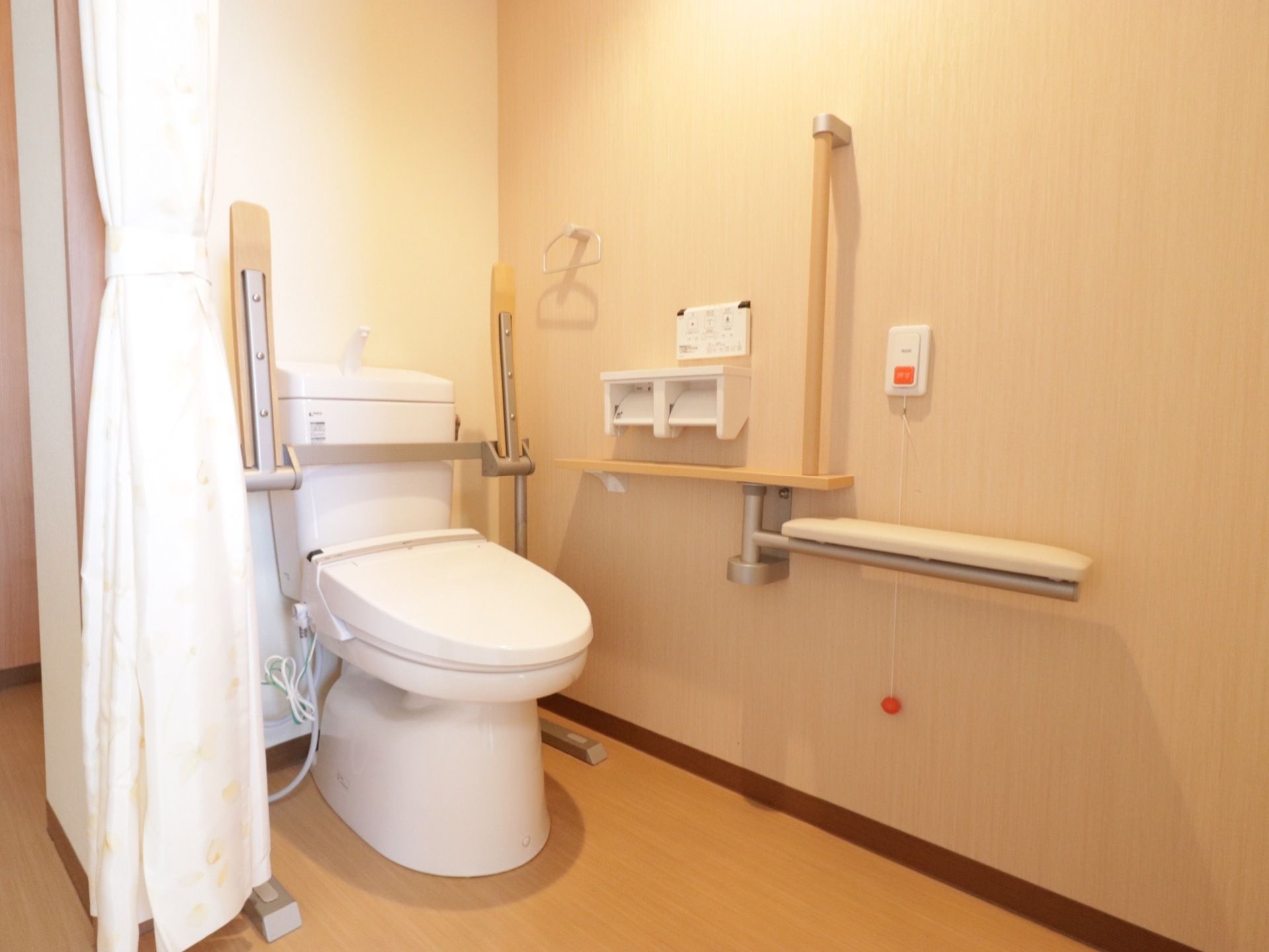 トイレの扉はカーテンタイプになっており、便器の脇には可動式の手すりが設置されている。壁にはナースコールが取り付けられている。
