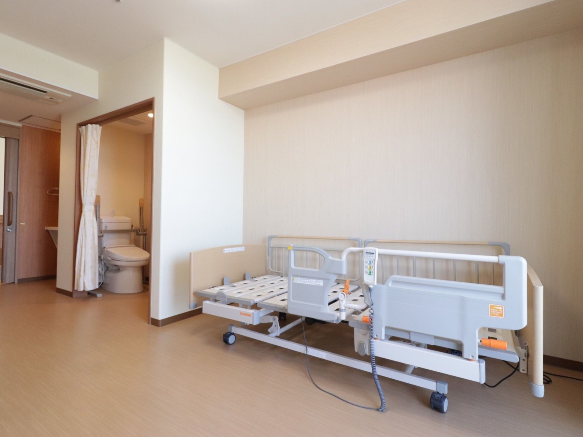 ライトベージュの壁とベージュの床が明るい雰囲気の居室。フロアには電動式ベッドが置かれている。トイレの扉はカーテン式になっている。