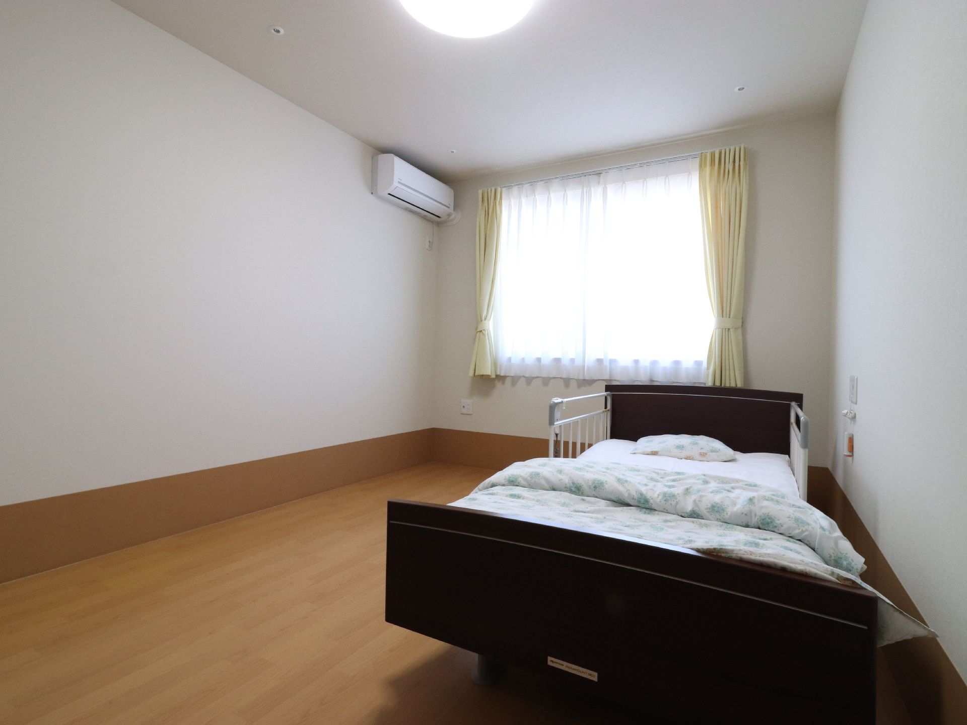 居室の床は、フローリングになっている。ベッドには、頭部両脇に手すりがついている。壁と天井は白色、床は茶色で統一されている。