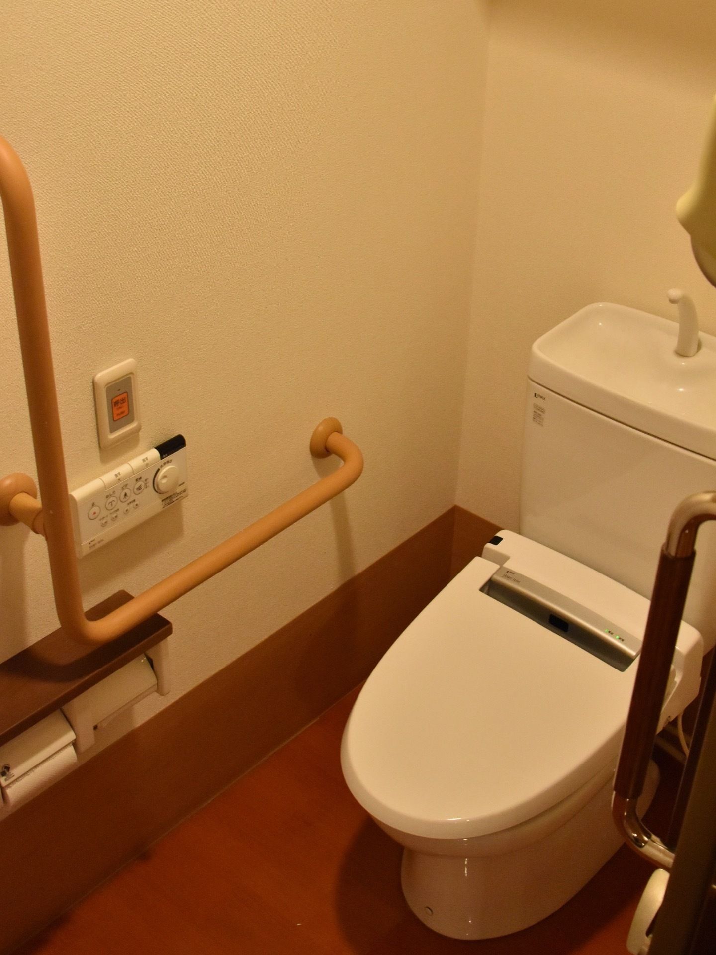 居室内にはトイレがあり、温水便座、Ｌ字型手すり、緊急呼出しボタンが設置されている。フローリングの落ち着いた内装である。