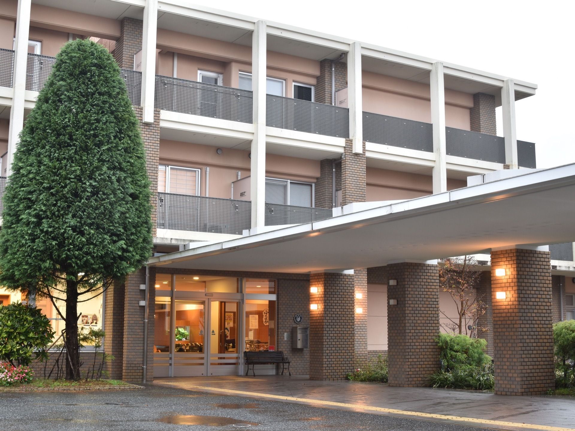 ３階建の建物。茶色系の温かみのある外観である。玄関の周りにはたくさんの樹木が植えられている。玄関のアプローチには屋根がかかっている。