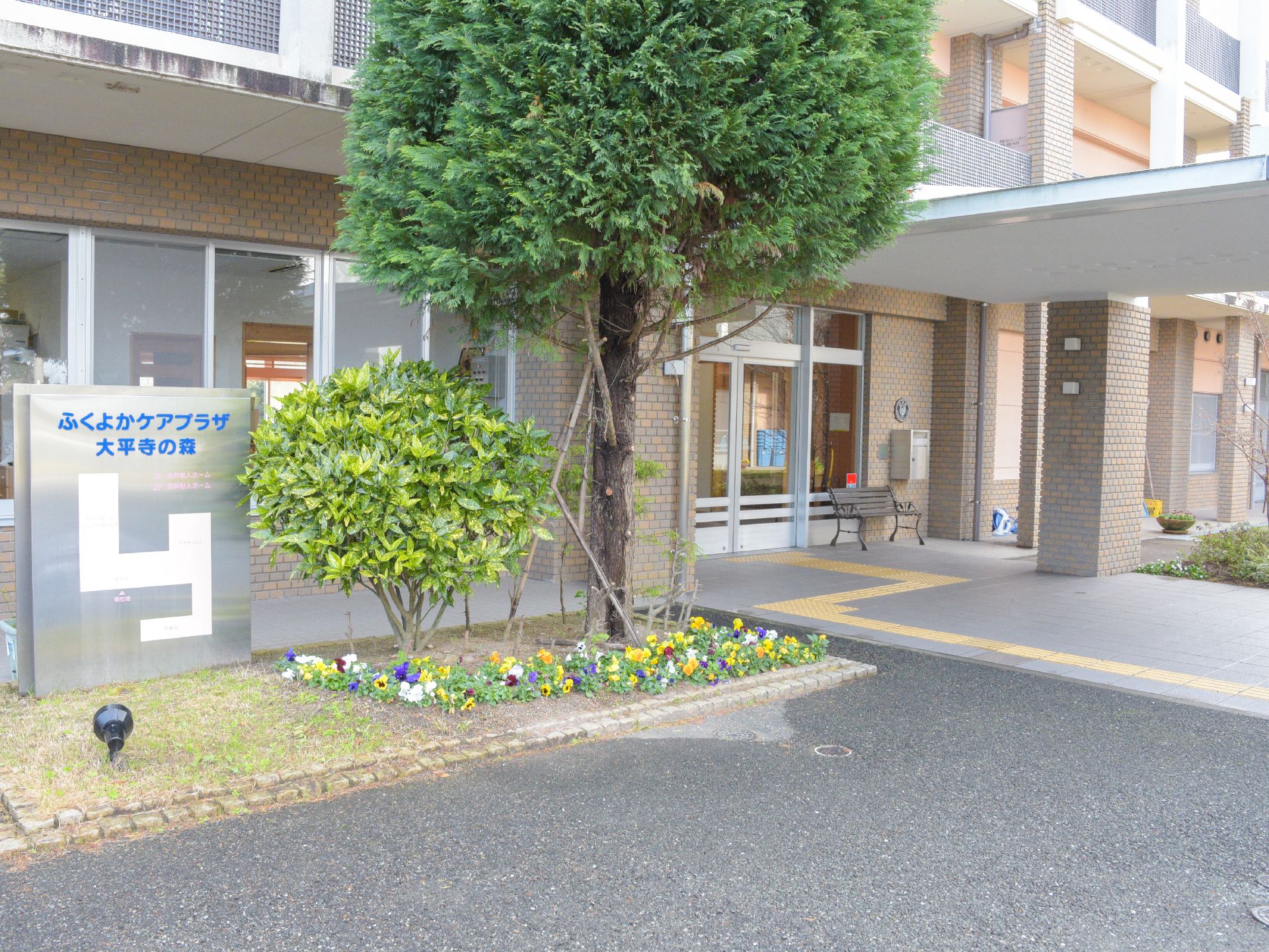 施設の前には芝生があり、花が植えてある。低木が植えてある。玄関アプローチにはベンチ、ポストが置いてある。