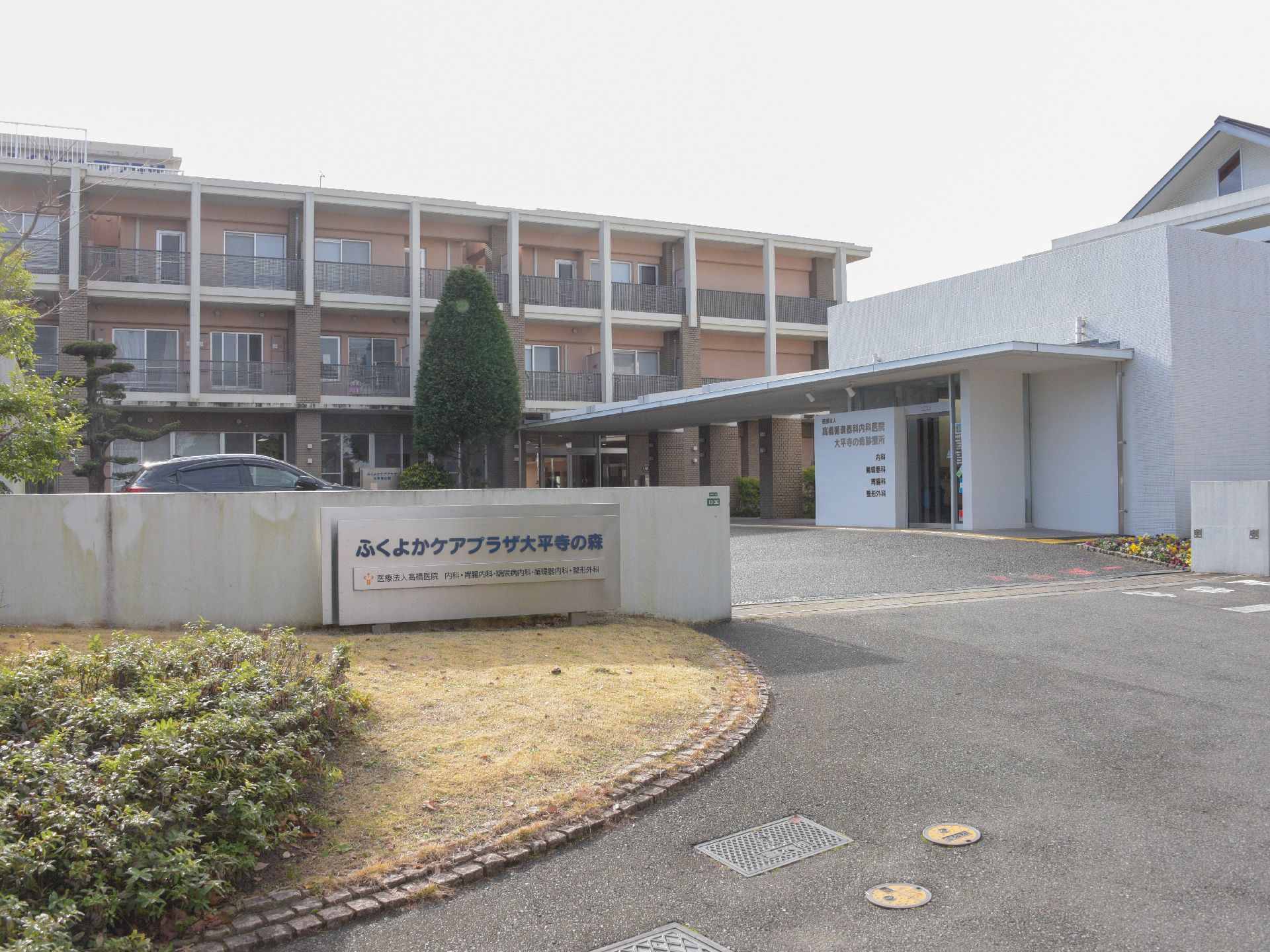 施設の前には塀があり、シルバーの表札がある。玄関アプローチには、長い庇の対らプロムナードがある。塀前に花壇がある。