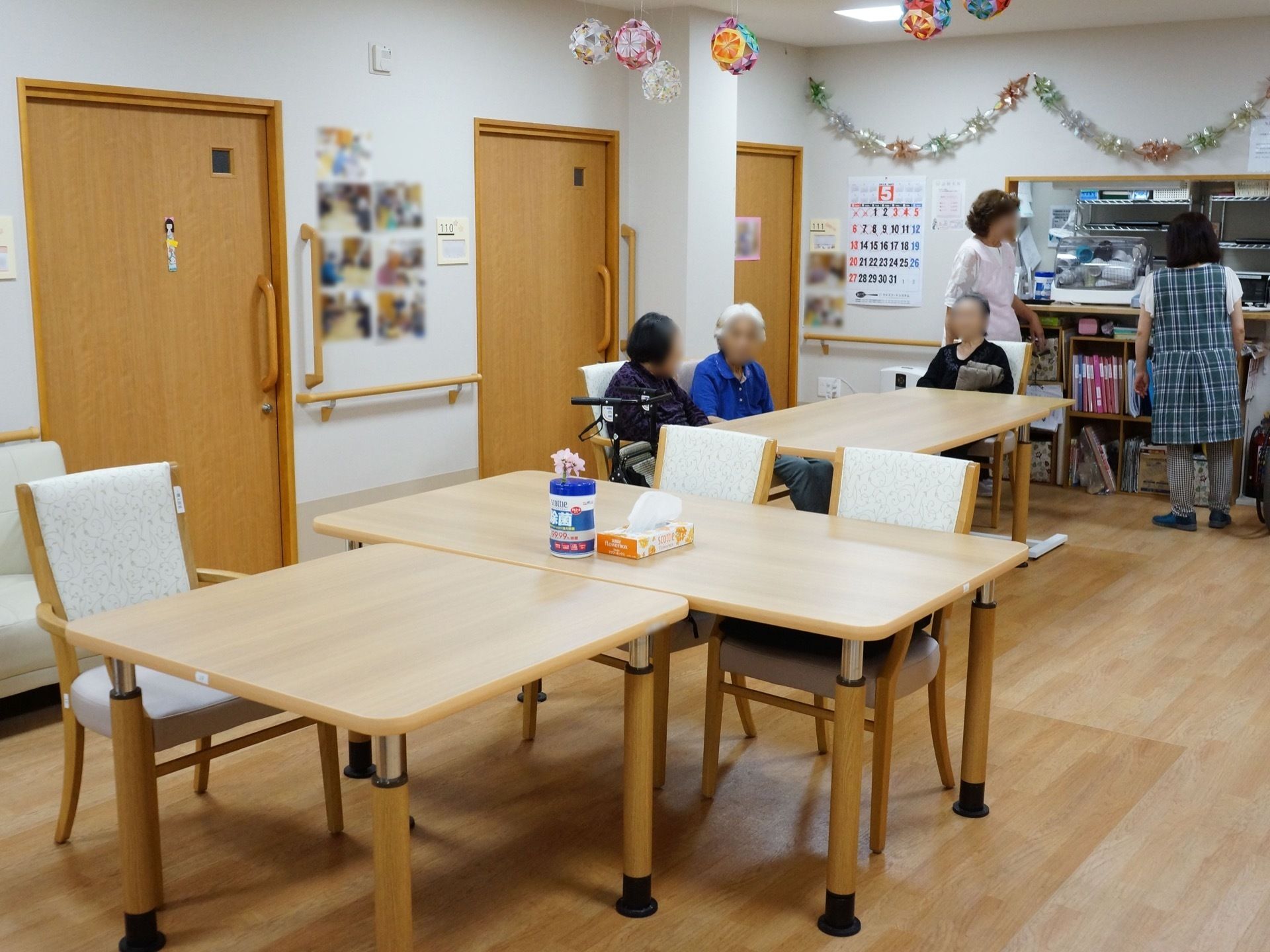 明るい木目調を基調にした食堂である。テーブルセットが設置されている。３名の女性の入居者様が、テーブルの椅子に腰掛け雑談されている。