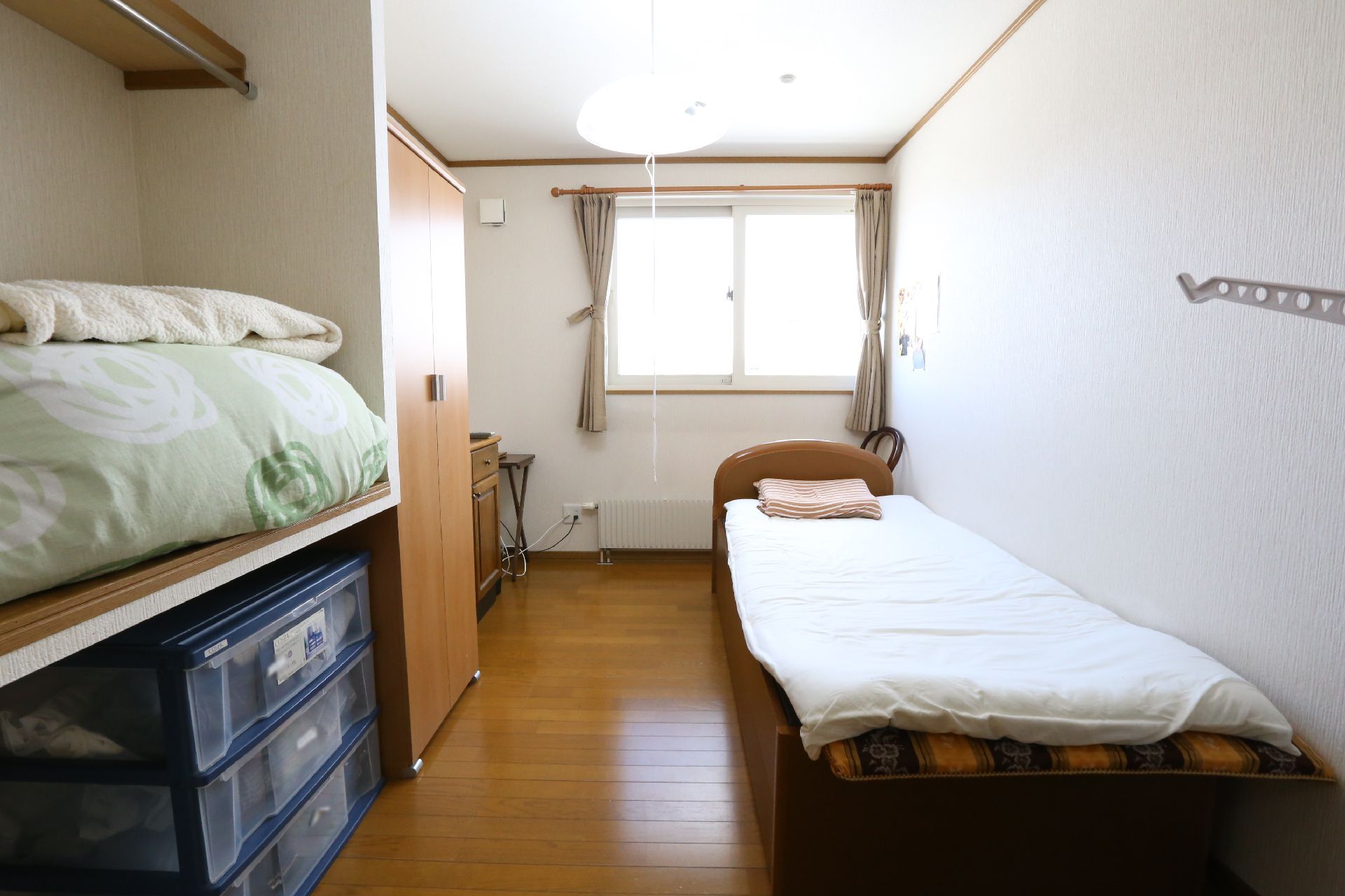 壁側にベッドが設置されている。天井に丸型のライトがあり、紐がベッド近くまで伸びている。窓側に椅子が置かれている。