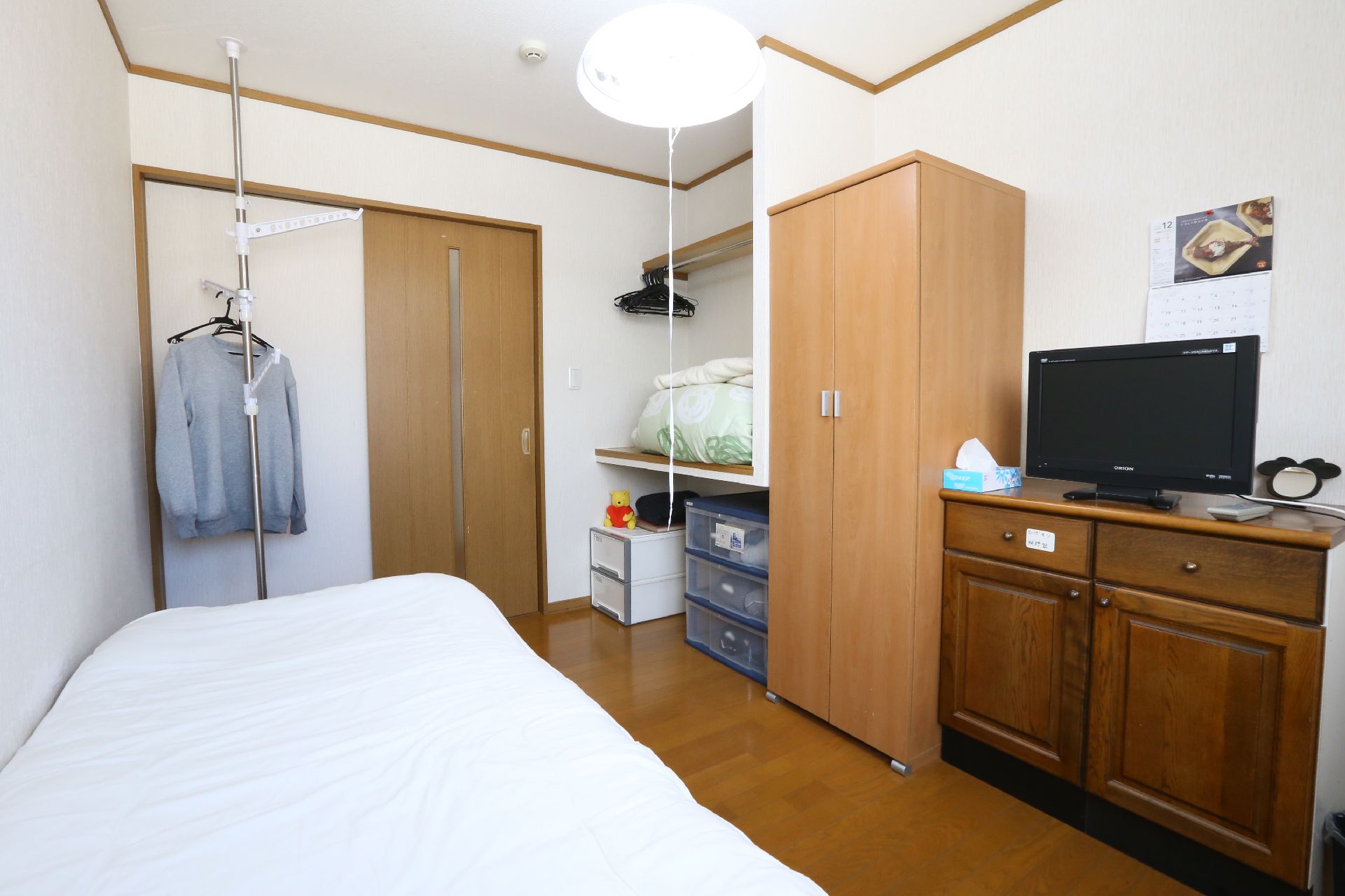 ドアの近くに備えつけの収納棚がある。壁側にベッドが置かれている。茶箪笥の上にテレビが設置されている。