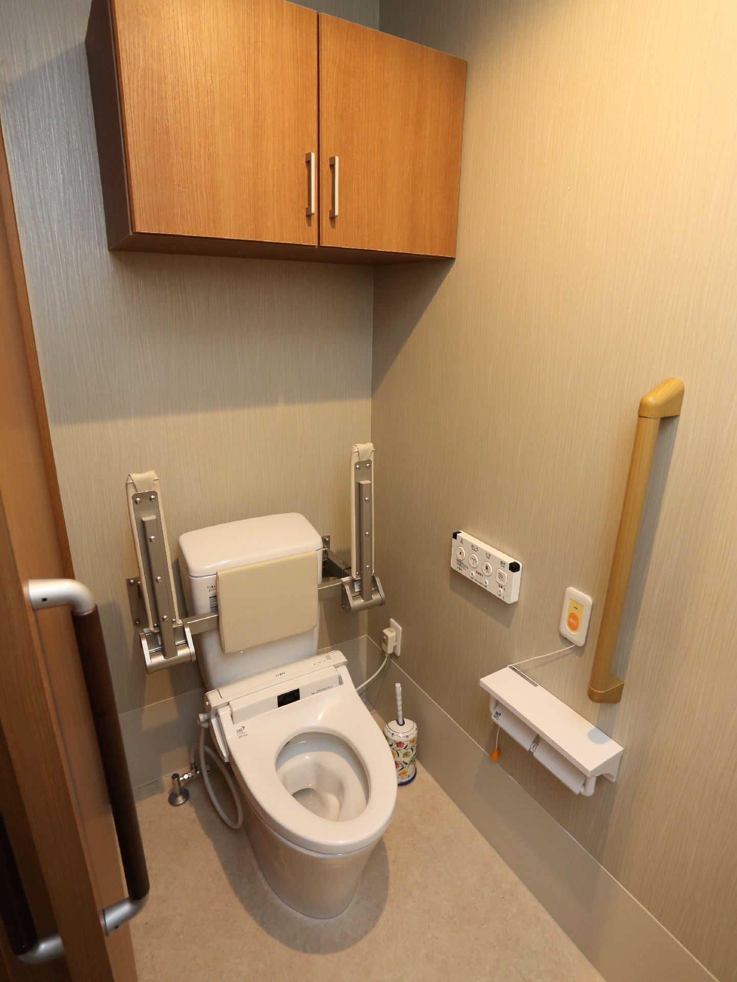 施設利用者のトイレである。両脇に肘置きと背もたれが取り付けられている。また壁には手すりや緊急呼び出しボタン、上部に収納棚が設置されている。