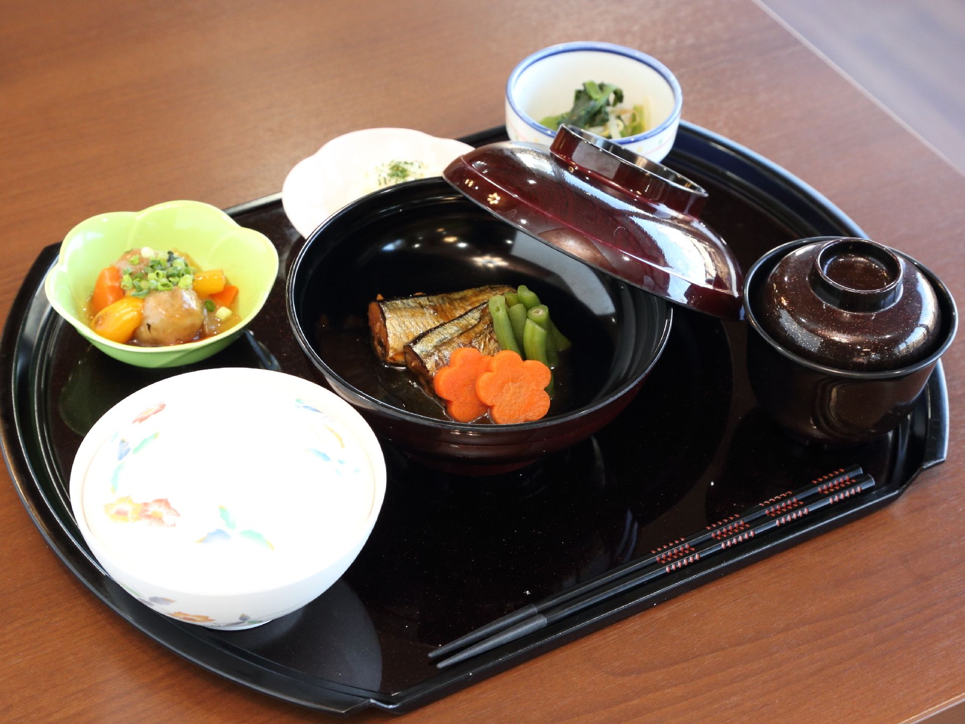 施設で提供される食事の一例である。魚のメイン料理に小鉢が3つ、ご飯とみそ汁がある。野菜が多めの和食メニューだ。