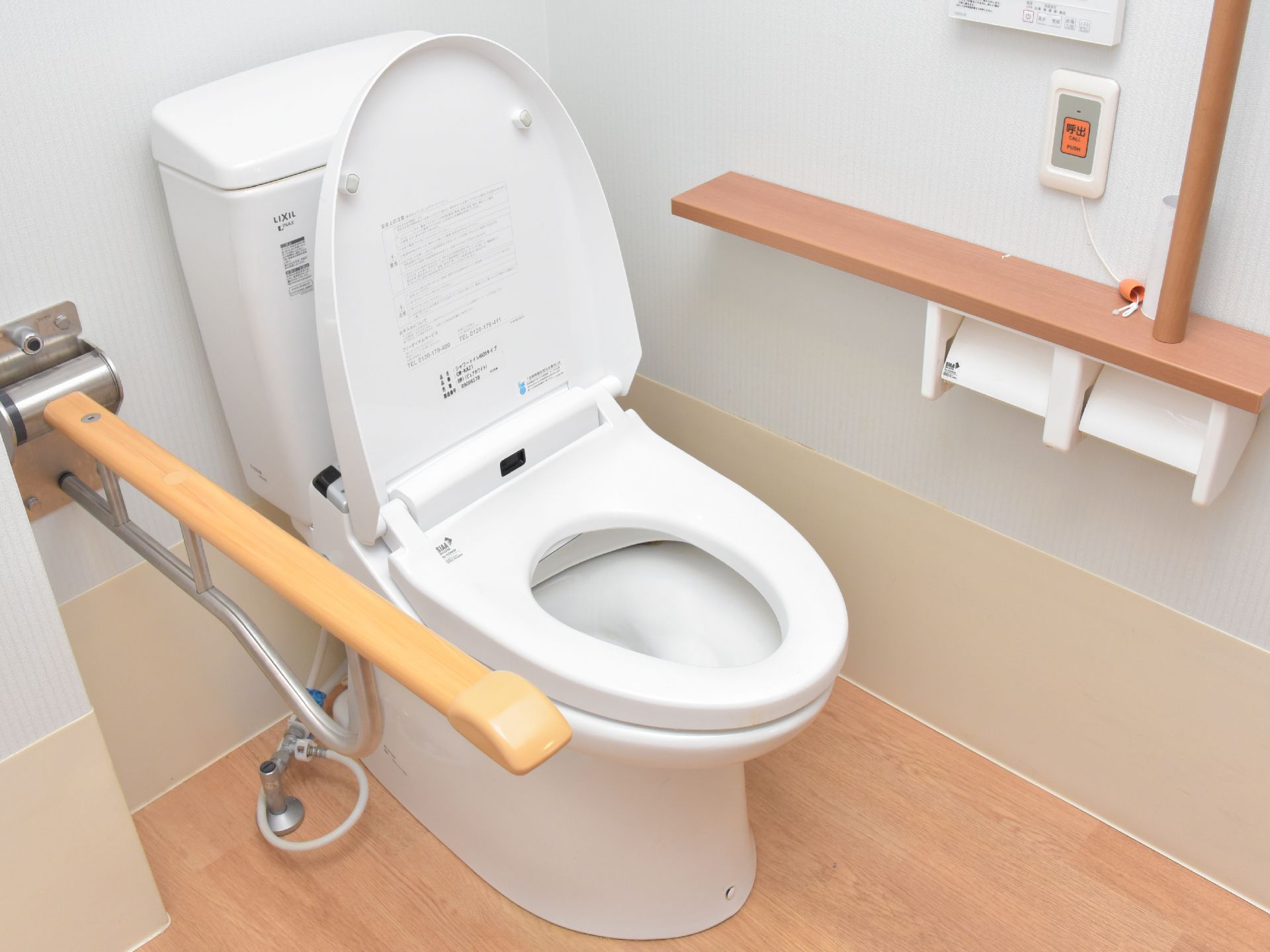 緊急時呼び出し用のブザーが壁に設置してある。ウォシュレット用のリモコンパネルが壁にある。手すりがついた洋式トイレである。