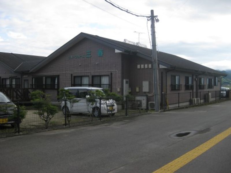 介護施設の建物外観。茶色いタイル張りの平屋建て施設で、駐車場と車が見える。