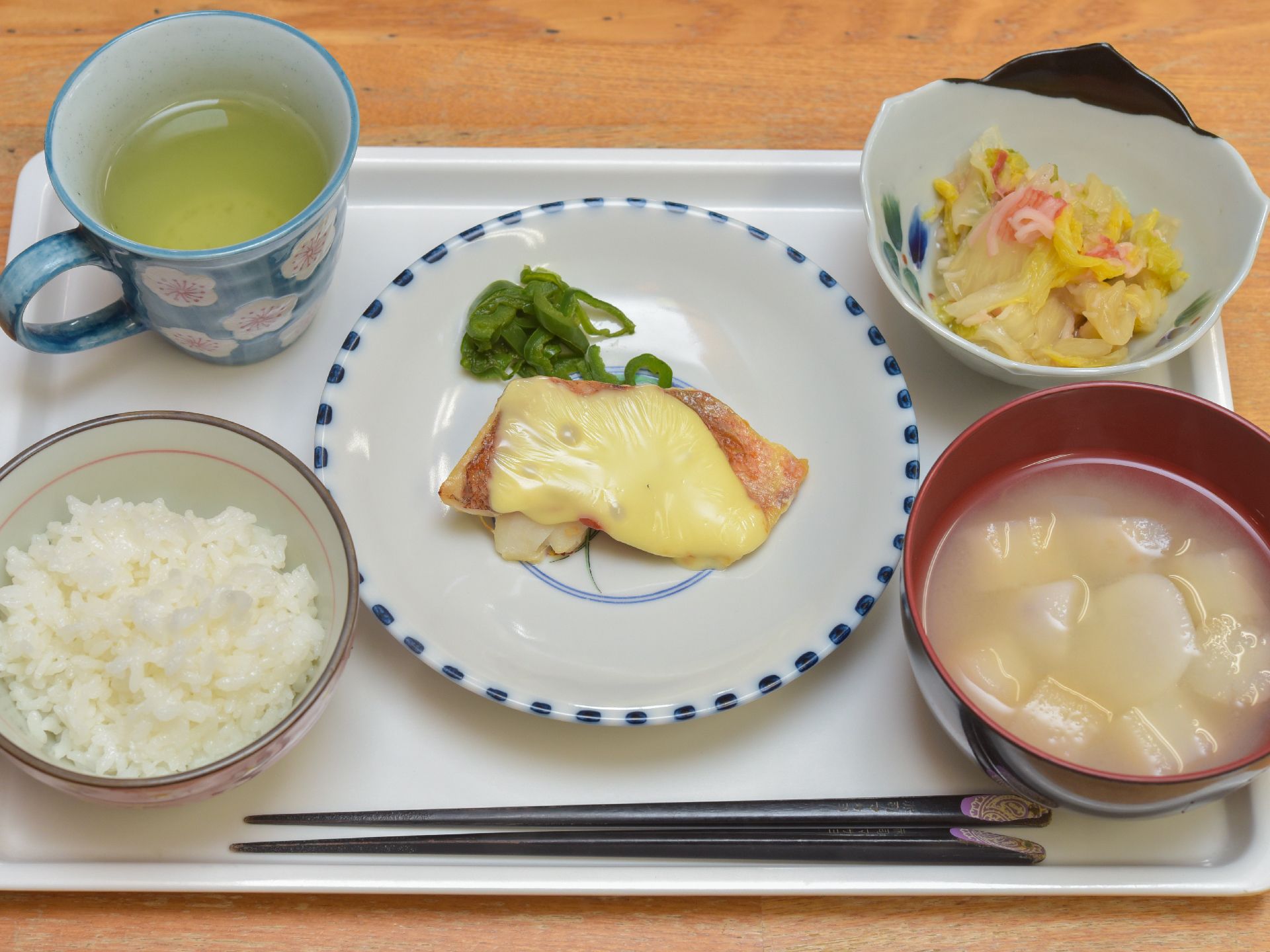 木製のテーブルの上に、白いトレーが置いてある。ごはん、味噌汁、小鉢が1つ、丸皿1つが付いている食事である。お茶もついている。