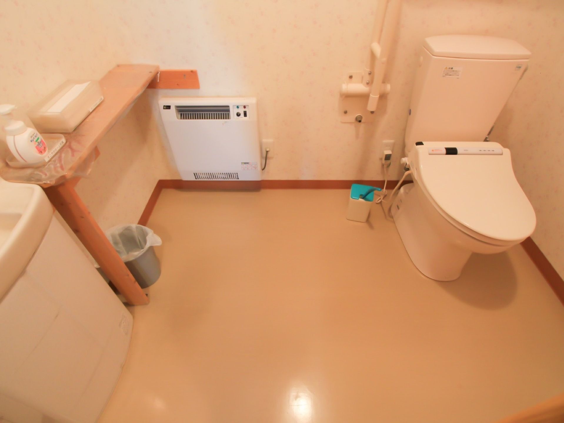 広いトイレである。暖房が完備されている。便座横にははね上げ式の手すりが設置されている。ハンドソープ、ペーパータオルも置いてある。