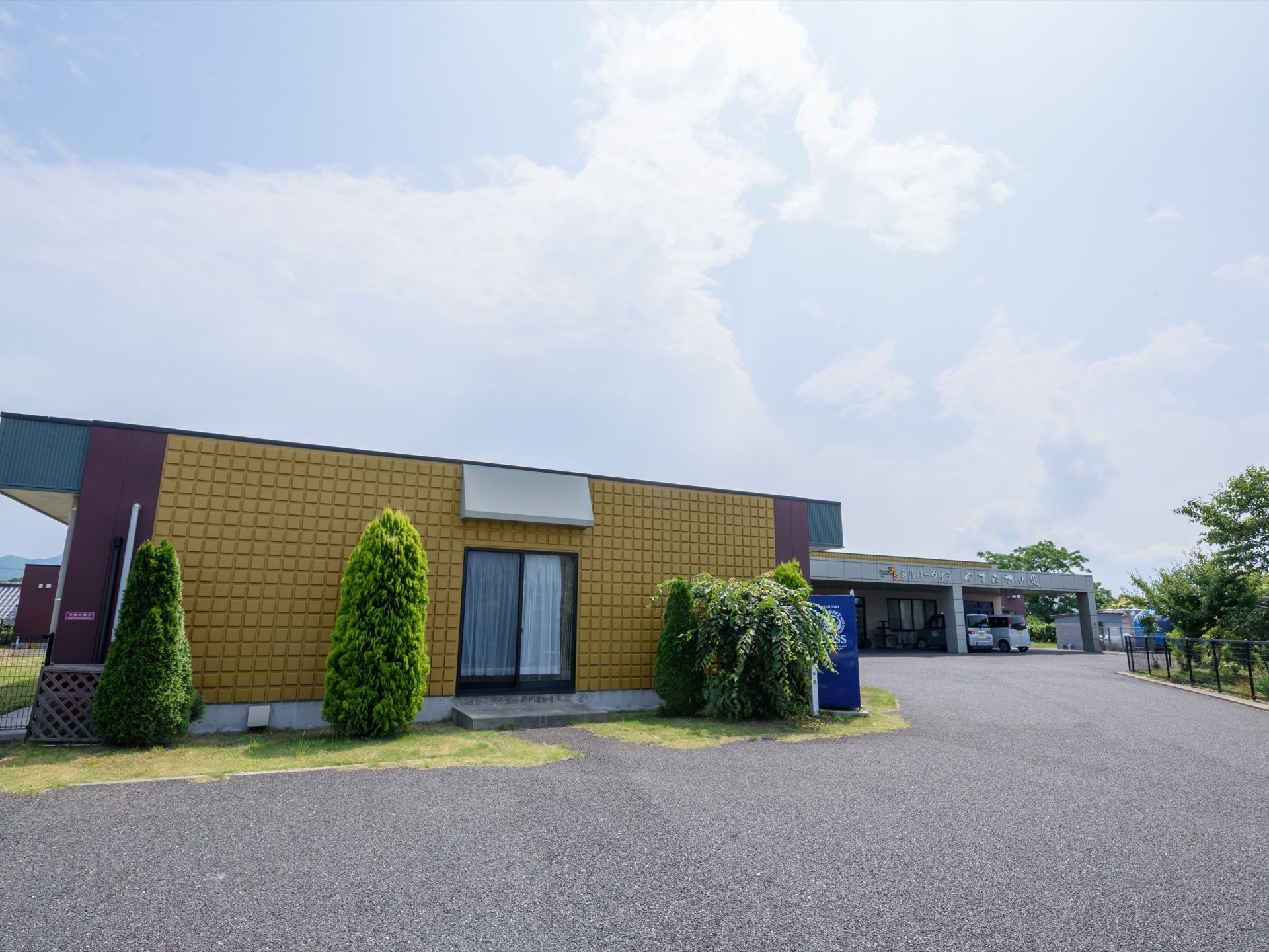施設の外壁は黄色く、目立っている。窓があり、窓付近には3本の木が植えられている。自動販売機が設置されている。