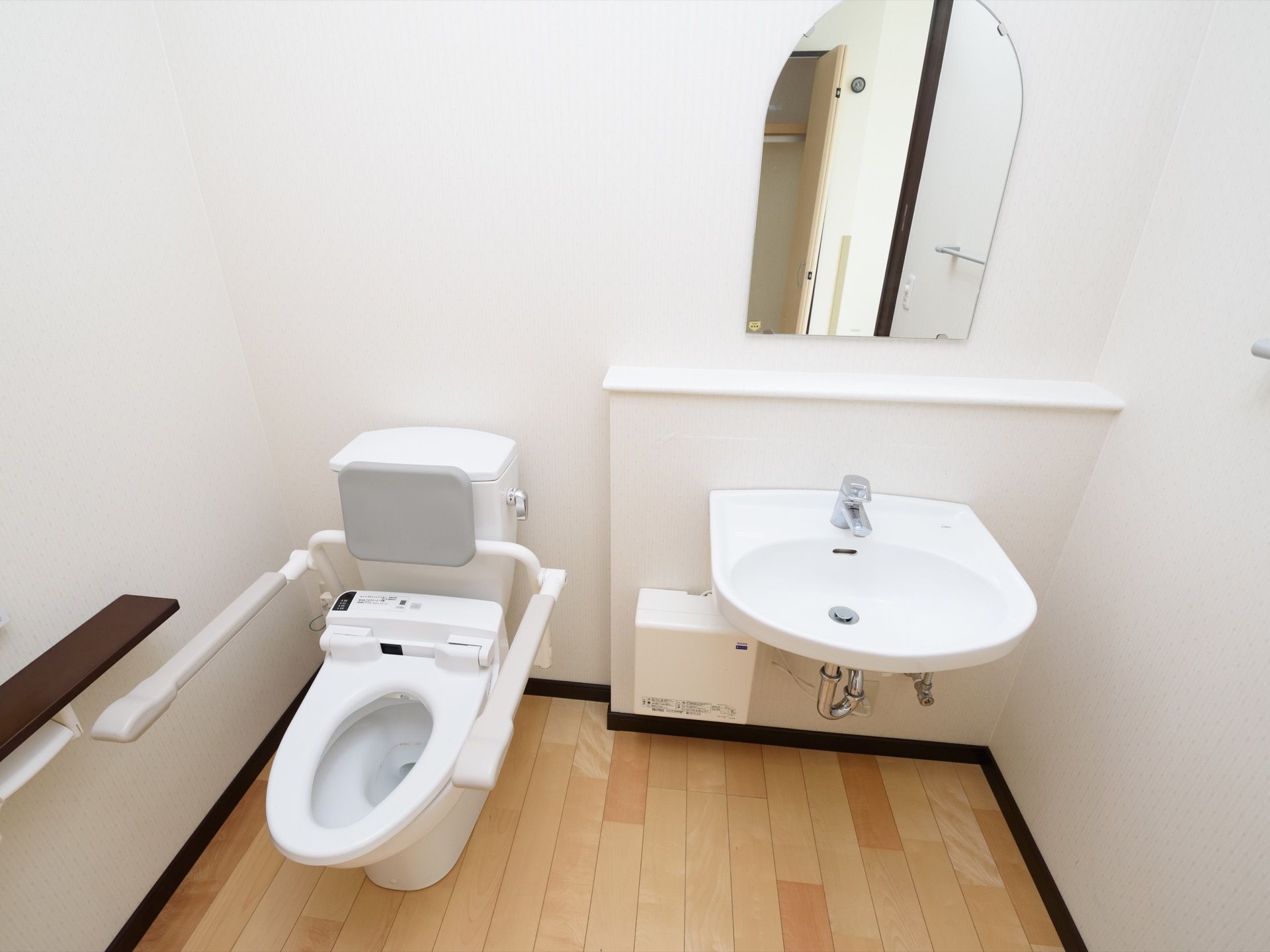 トイレには洋式便器、手洗い器が同じスペースにある。便器横には可動式の手すり、壁側のペーパーフォルダーの上には棚がある。