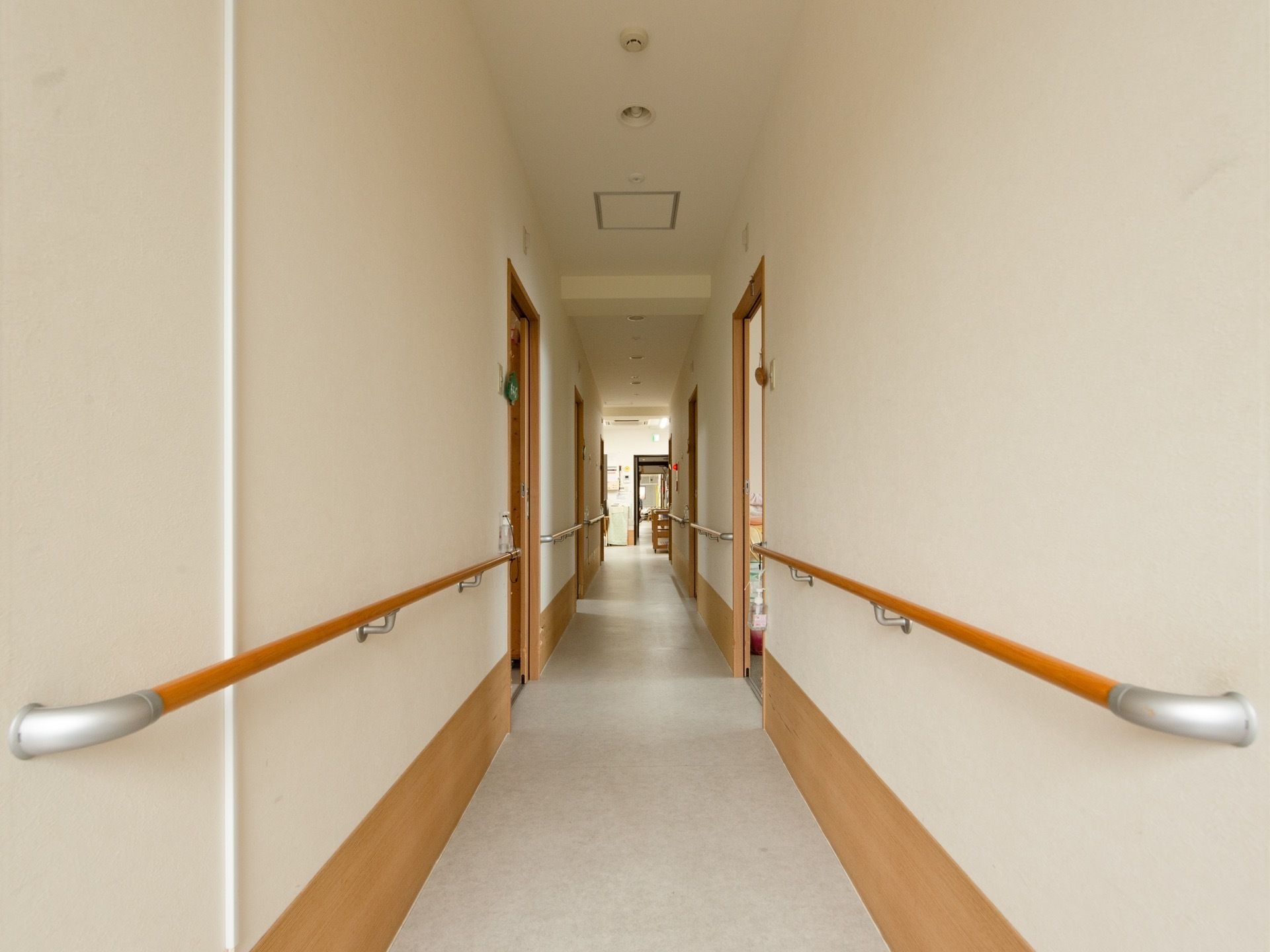白い壁とグレーの床の長い廊下がある。両サイドには居室のドアが並んでおり、壁には手すりが設置されている。