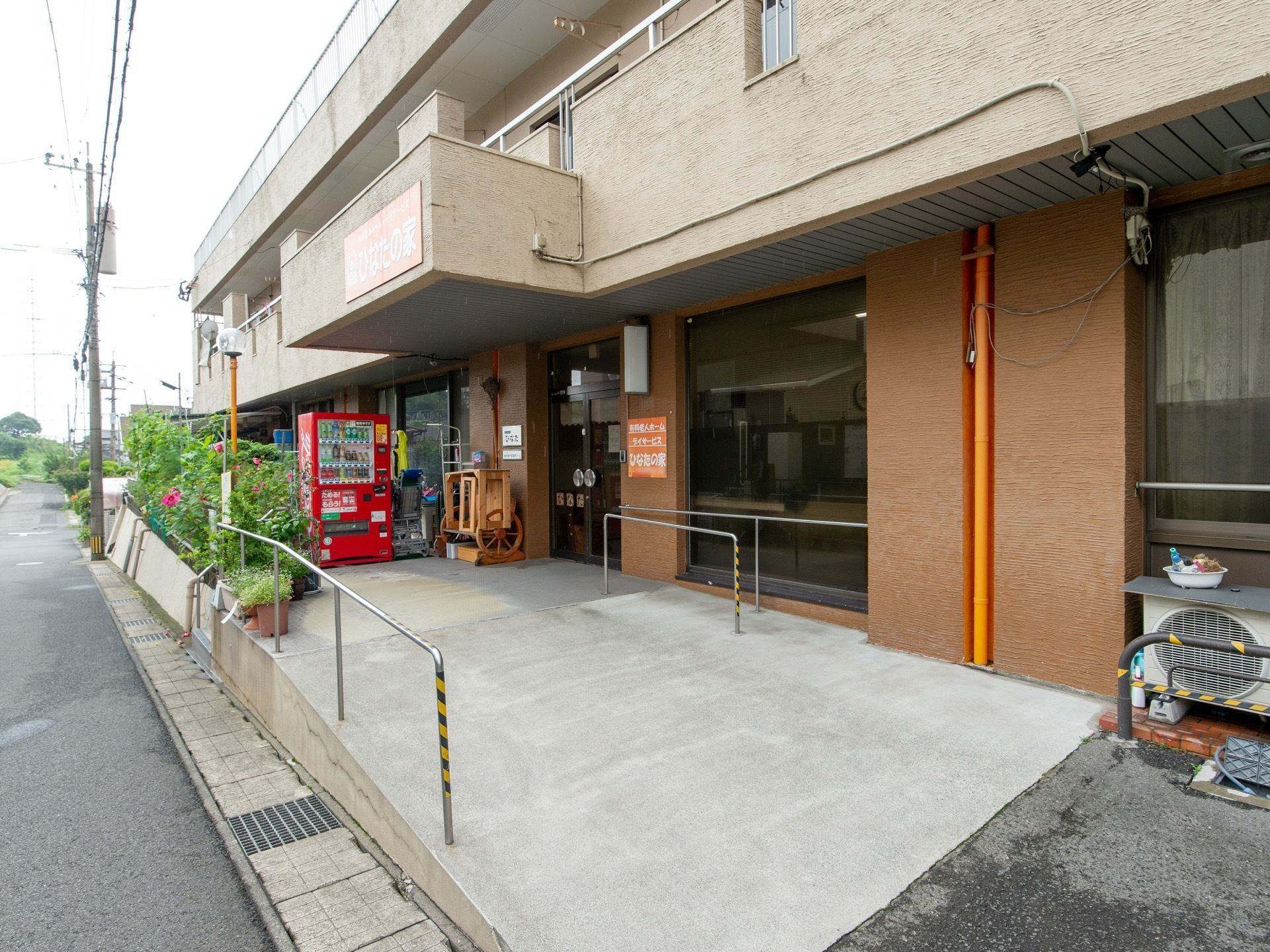 建物の玄関に向かって幅の広いスロープが設置されている。両側には手すりがある。玄関前には植物が生い茂り、自動販売機が設置されている。