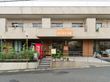 坂道に面した２階建ての建物。一階部分はガラス張りになっており、玄関ドアに向かってスロープと手すりがある。たくさんの植物が植えられている。