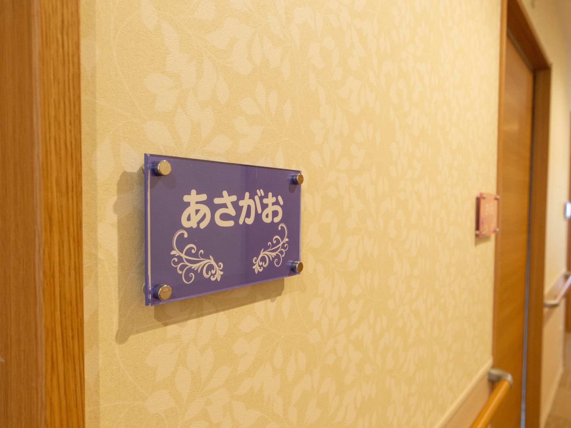 葉っぱの模様が描かれたベージュ色の壁に、花の名前が書かれたプレートが取り付けられている。木目調のドアが並んでいて、壁には手すりが付いている。