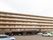 ベージュとブラウンの2色の外壁になっており、建物は6階建てになっている。手前は駐車場になっており、たくさんの車が停まっている。
