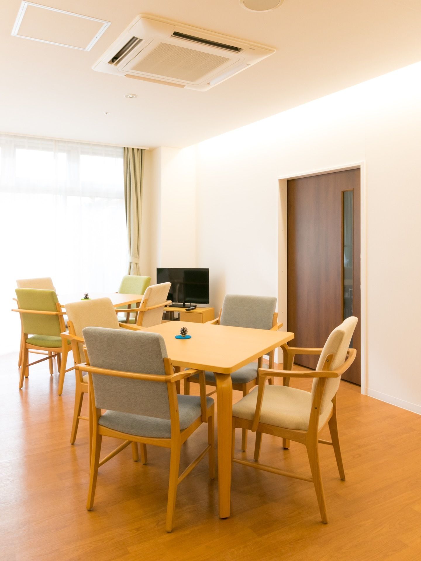 正方形のテーブルに、椅子が４脚ずつセットされている。部屋の奥にテレビが置かれている。ダークブラウンの引き戸がある。