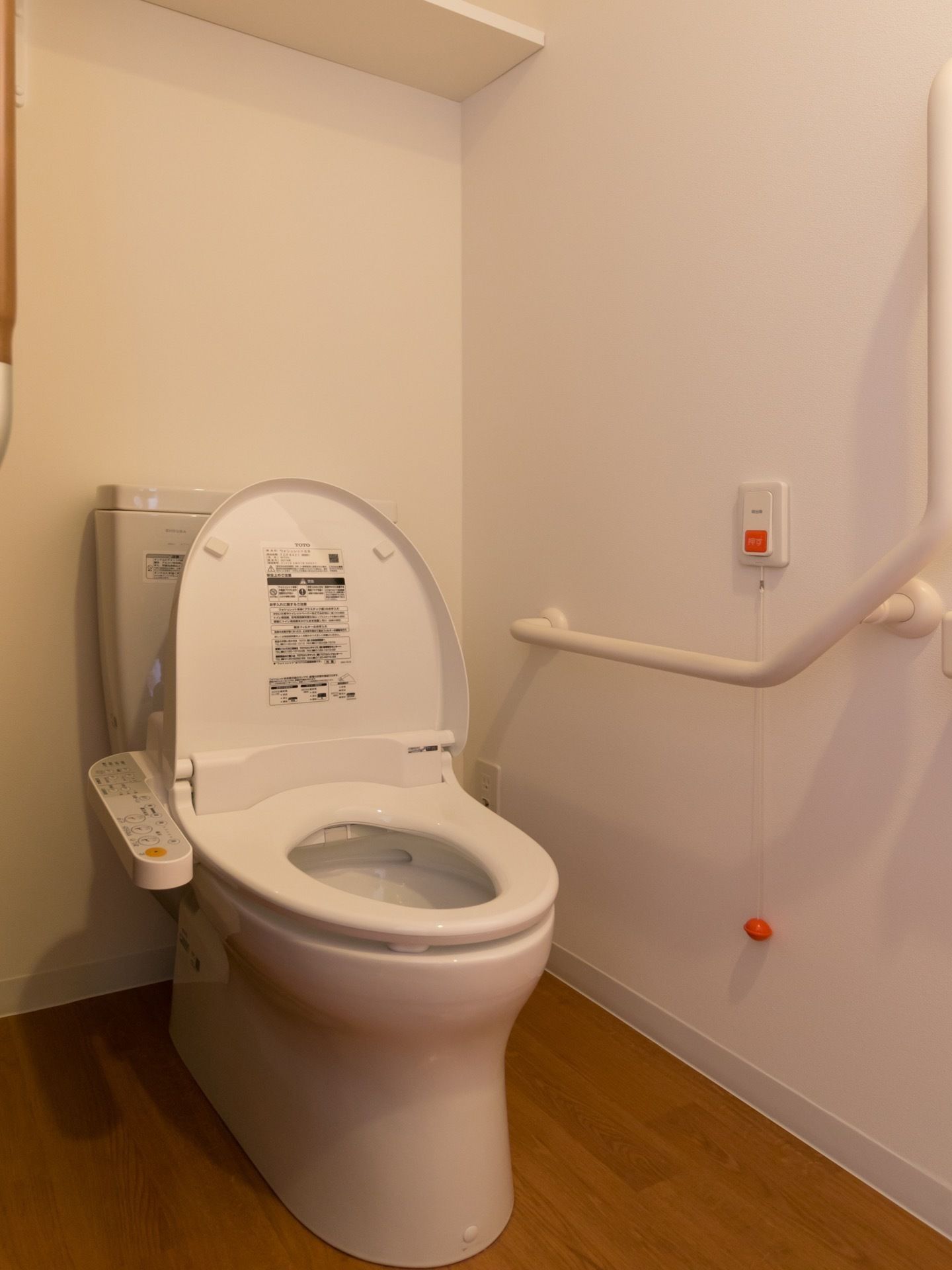 温水洗浄便座の付いたトイレである。壁には大きな手すりや、オレンジ色の緊急時の呼び出しブザーが設置されている。