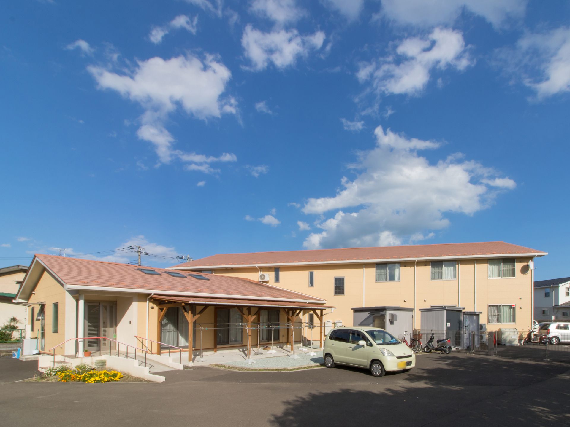 建物前に広い駐車場がある。入り口前は、手すり付きの階段とスロープがある。花の花壇がある。物干し台がある。