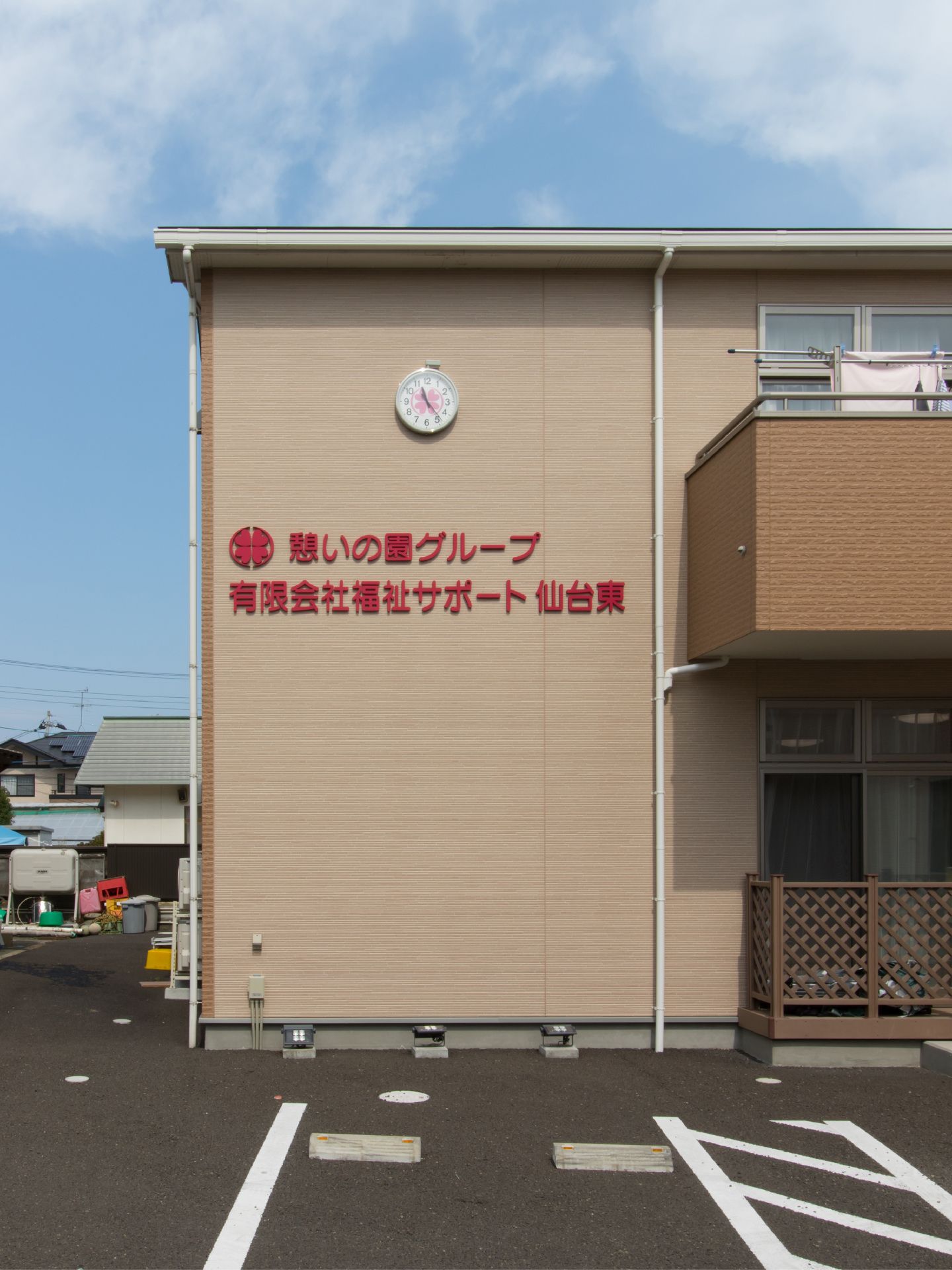 建物の外壁に、施設名が赤字で表示されている。時計が掛けられている。2階部分に洗濯物干しスペースがある。
