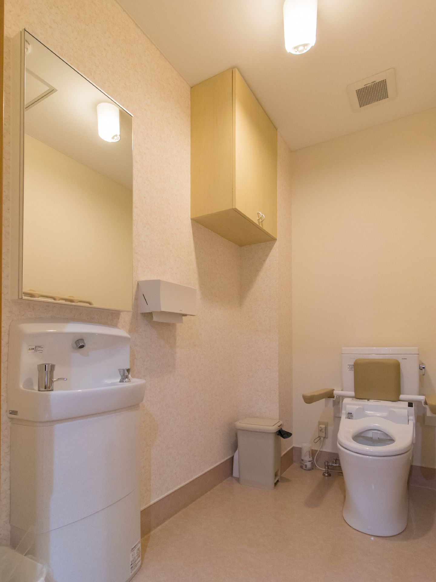 背もたれ付きの洋式トイレである。洗面所がある。鏡、ペーパーホルダー、ゴミ箱、収納棚、換気扇がある。壁と床は白色である。
