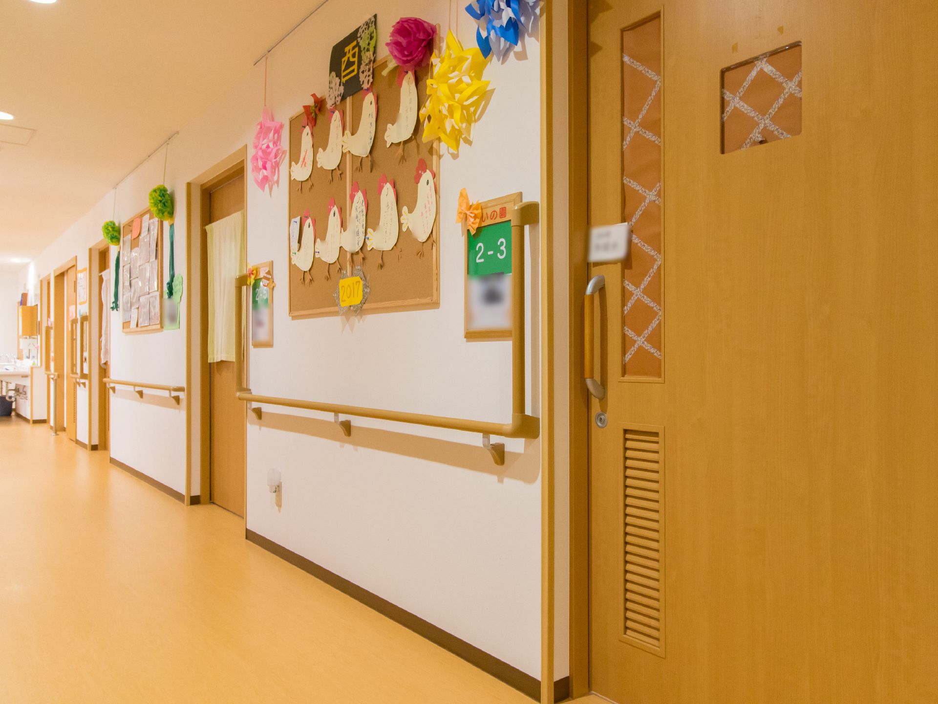 居室前に部屋番号と利用者様の写真が掛けられている。入り口ドアは引き戸式である。壁にはイベントで作成した作品などが飾られている。