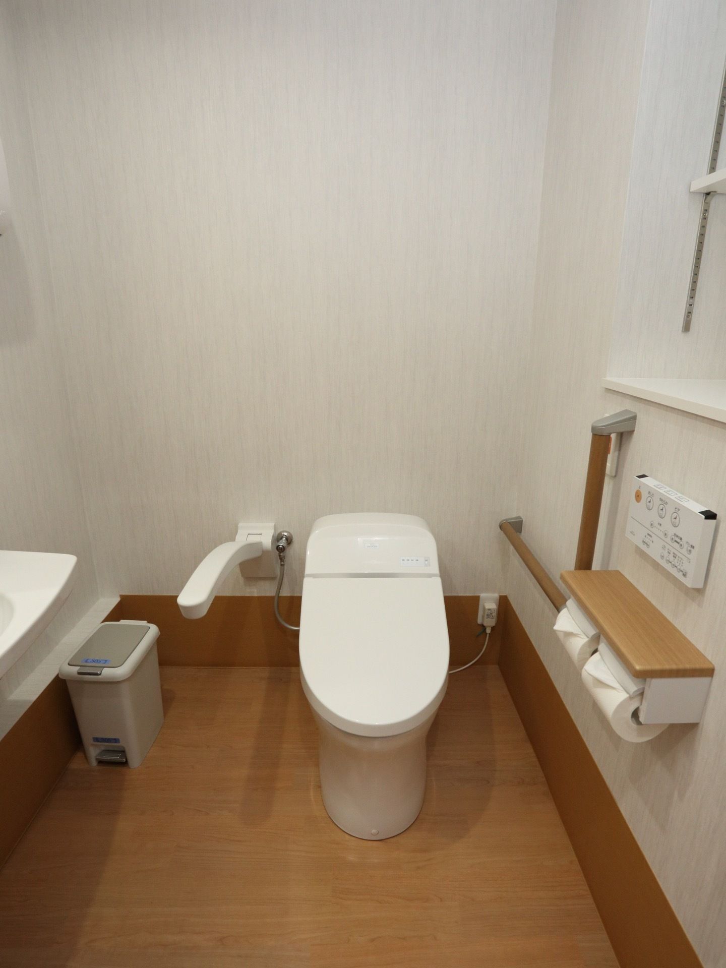 白と木目で統一され清潔感がある。コンパクトなトイレで周囲がスッキリしている。シャワーの操作盤が壁に付いている。
