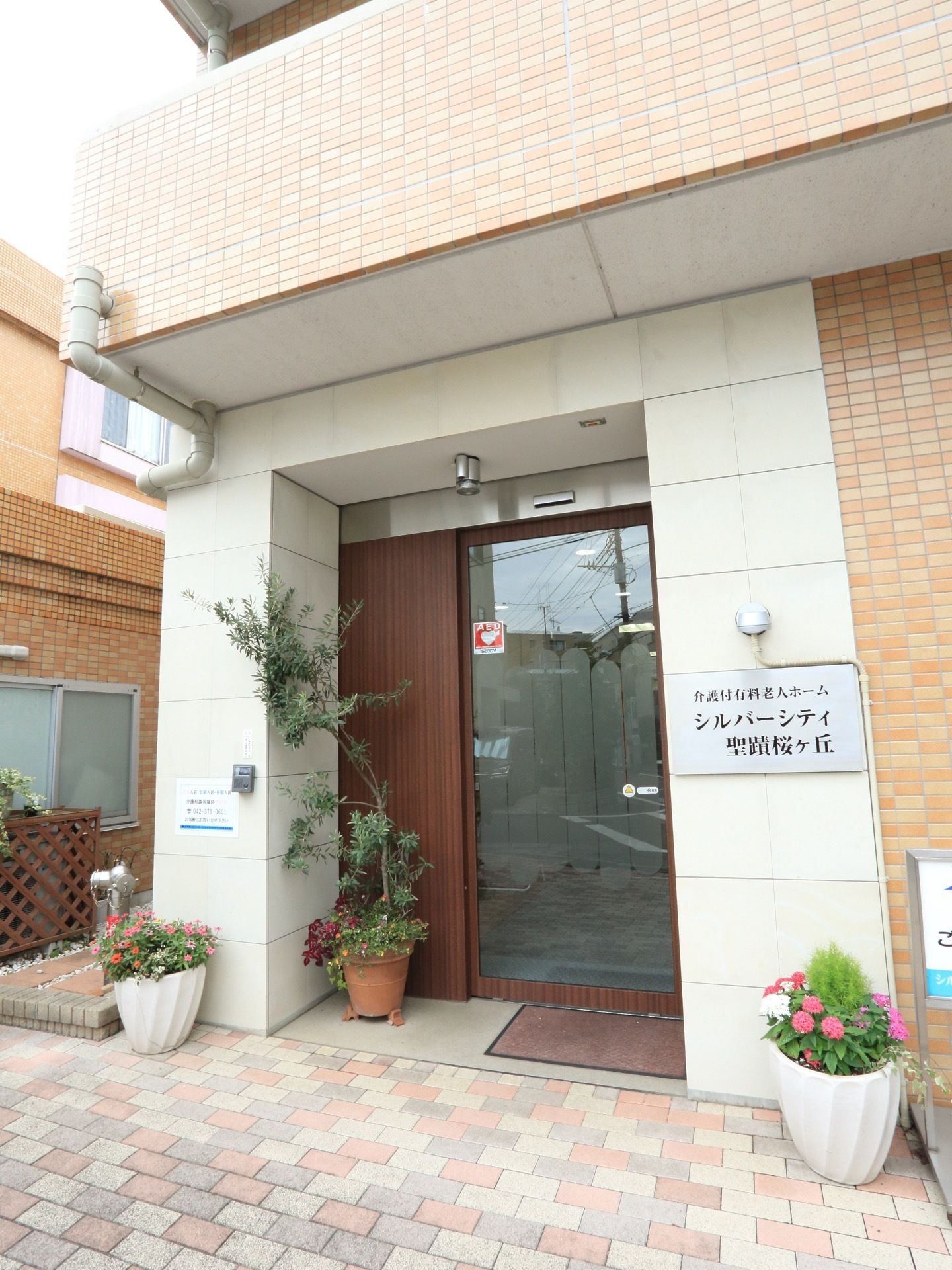 玄関アプローチはレンガ調のタイル敷きで、植木鉢が飾れている。玄関の壁には施設の表札がかかっていて、横には送水口が設置されている。