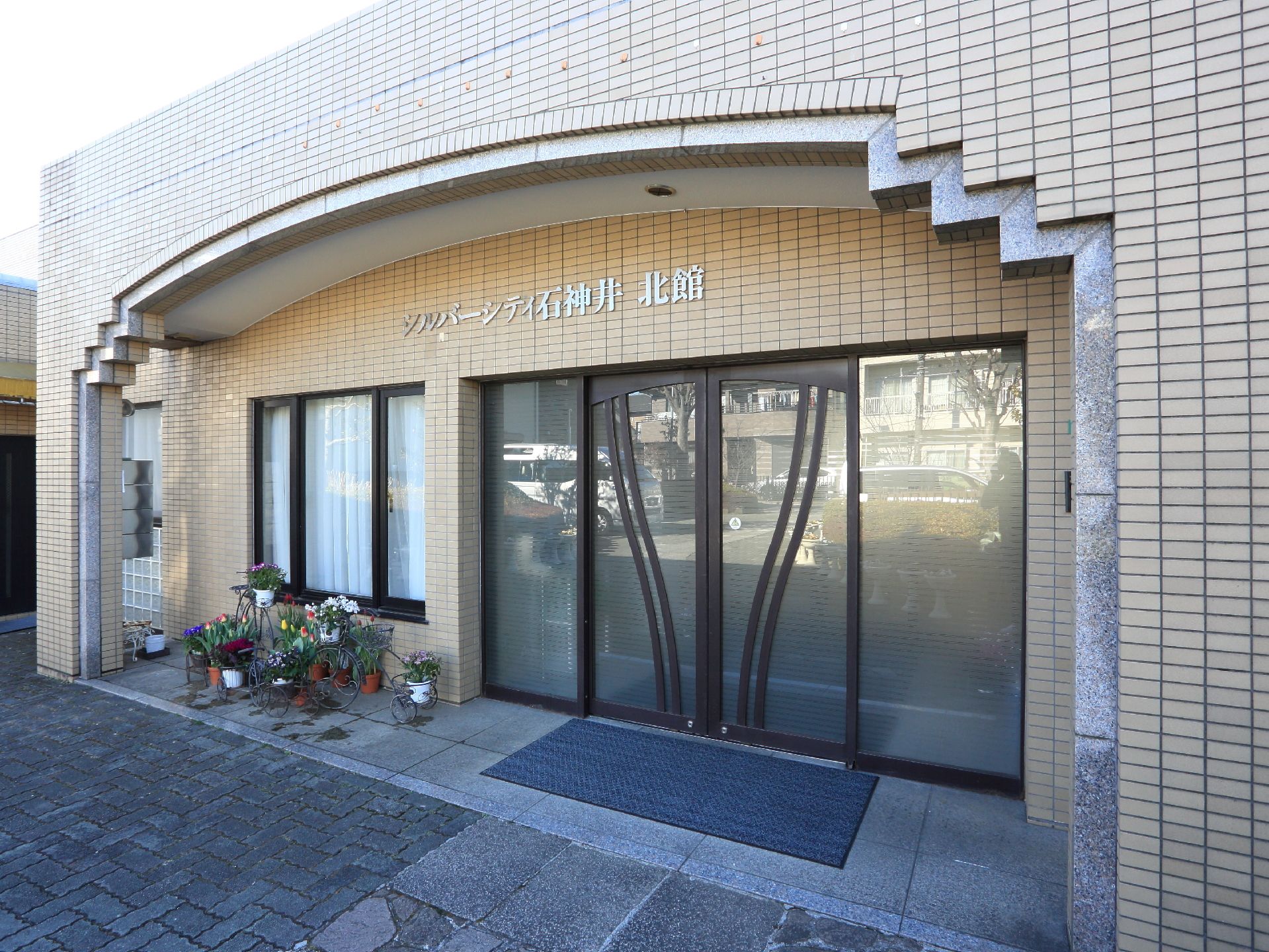 落ち着いた雰囲気の広いスペースの玄関である。入口の横には季節の花が咲いているプランターが複数置かれている。