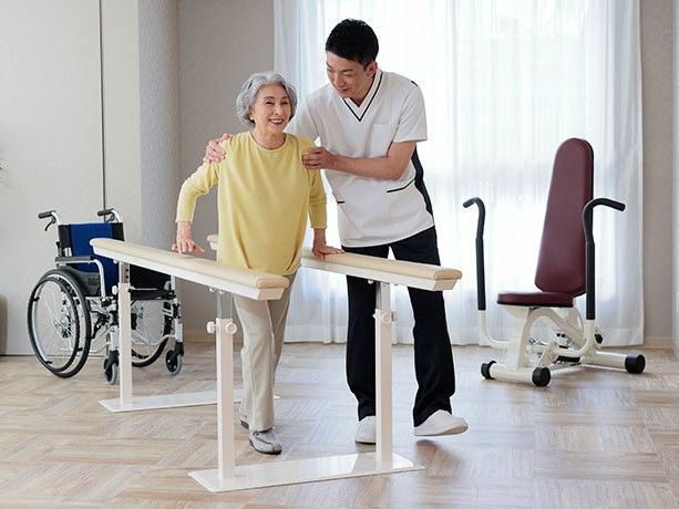 三人の高齢者がイスに座って、白い服のスタッフに運動らしきものを教わっている